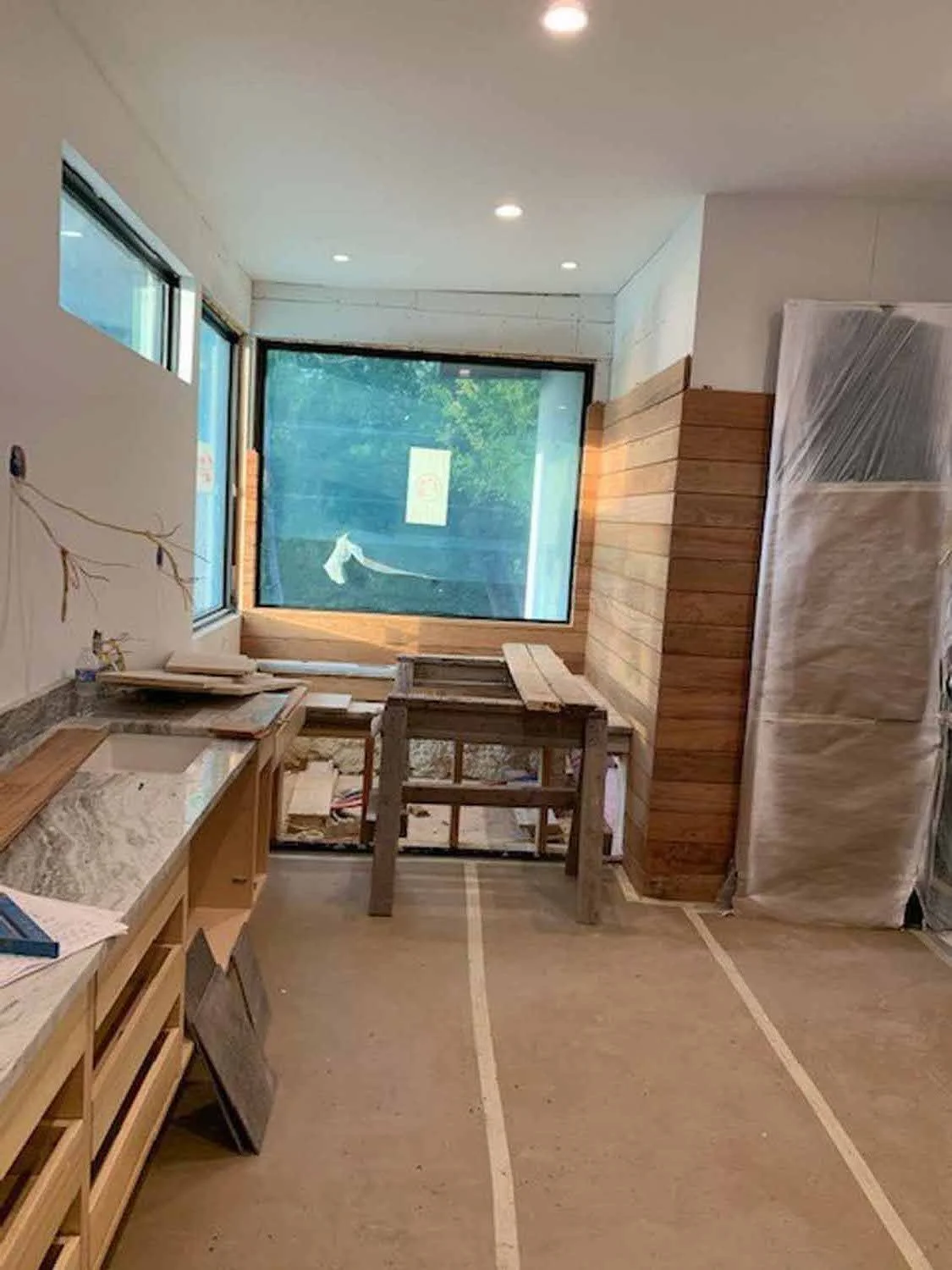 Interior kitchen under construction with wooden cabinets, a granite countertop, a large window, and construction tools and materials.