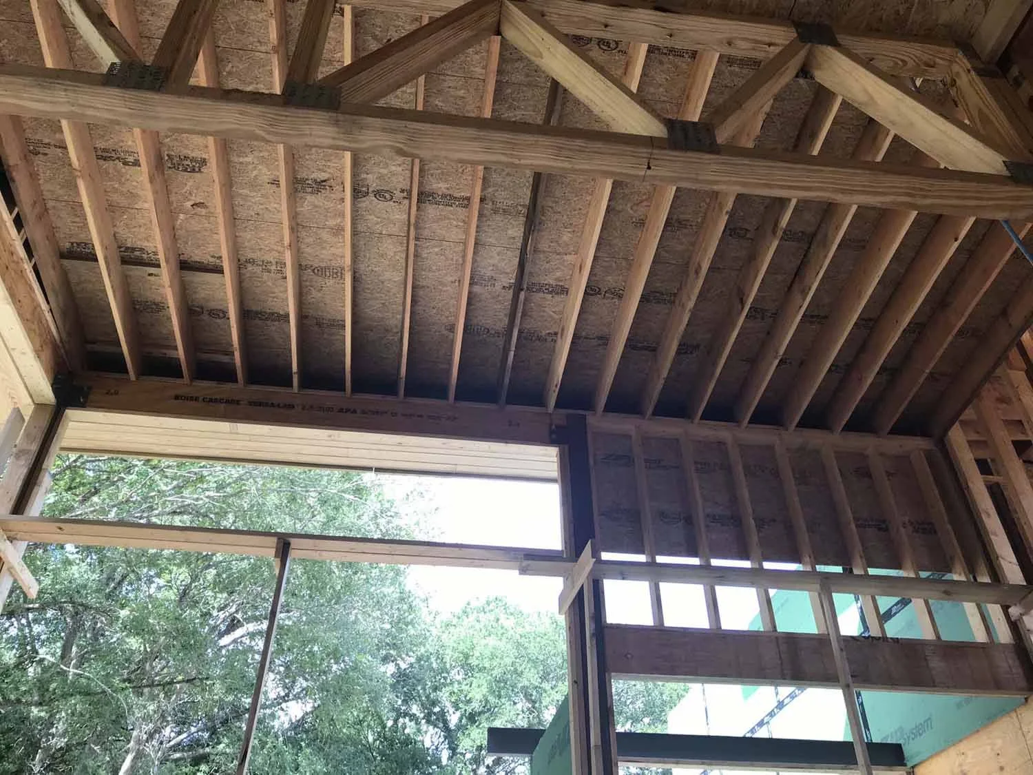 Interior of a building under construction with wooden framing and exposed ceiling, with a large opening showing outdoor trees.