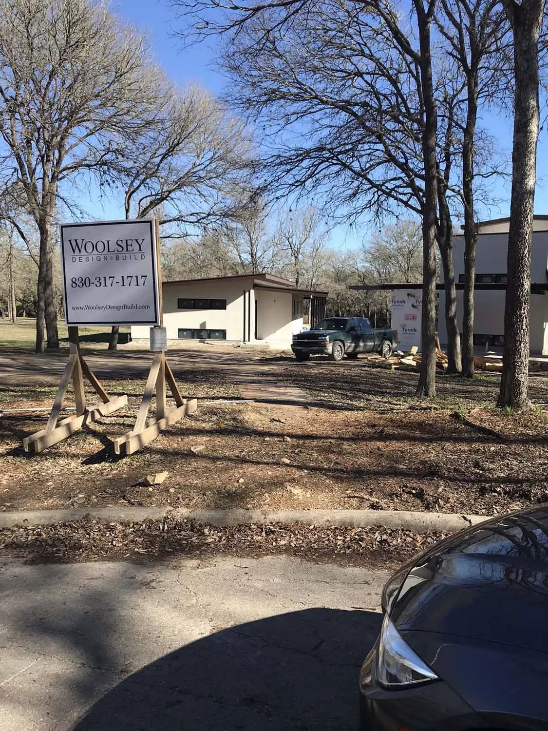 Construction site of a modern house with a sign that reads "Woolsey Design & Build" along with contact information, surrounded by trees and parked trucks.