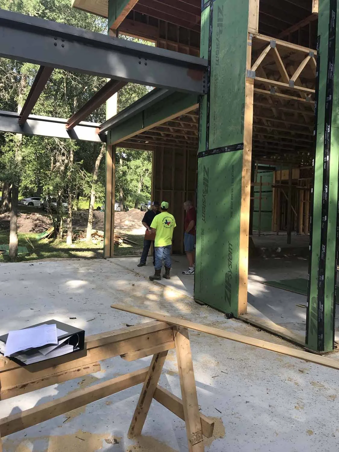 Construction site with workers discussing near the framework of a building, with exposed wooden and steel beams and green insulation panels, sunny day with trees in the background.
