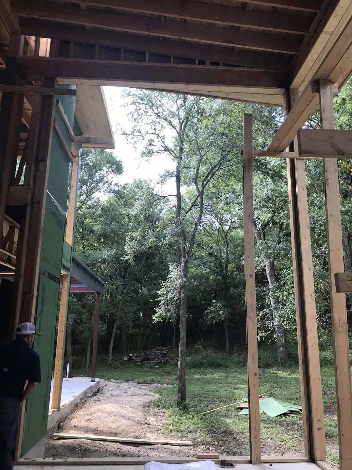 Construction site with wooden framing for a building, open view of trees and outdoors, a person with a cap looking at the construction.