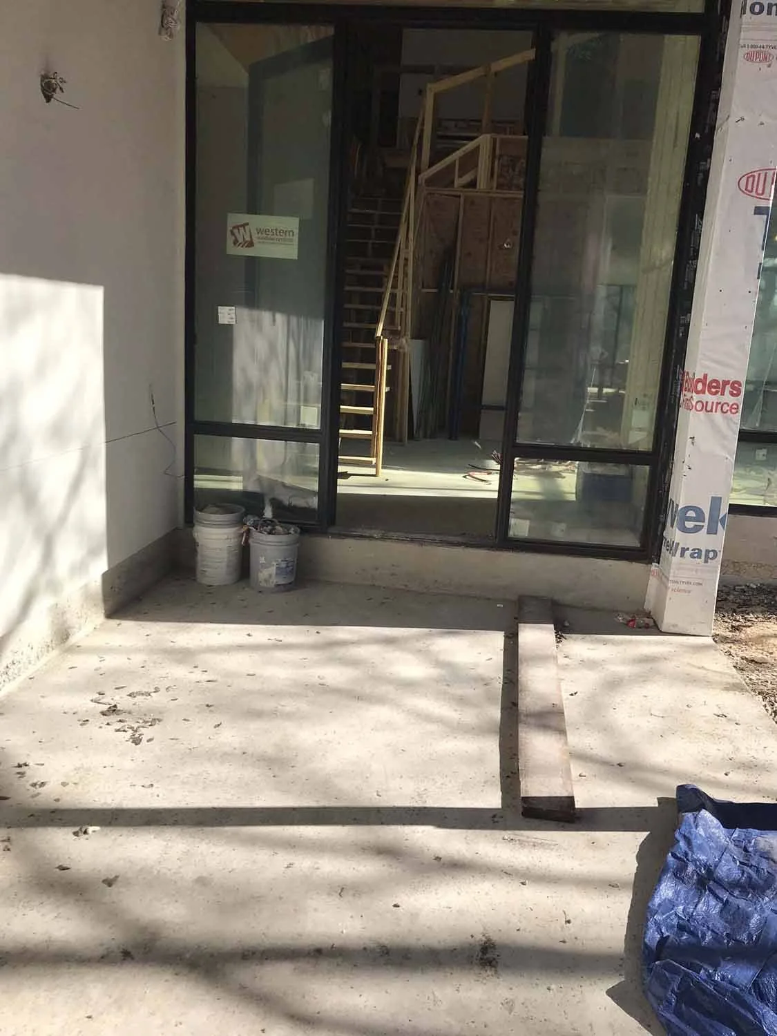 Construction site showing a building entrance with glass door and stairs inside, construction materials outside, and tools on the ground.