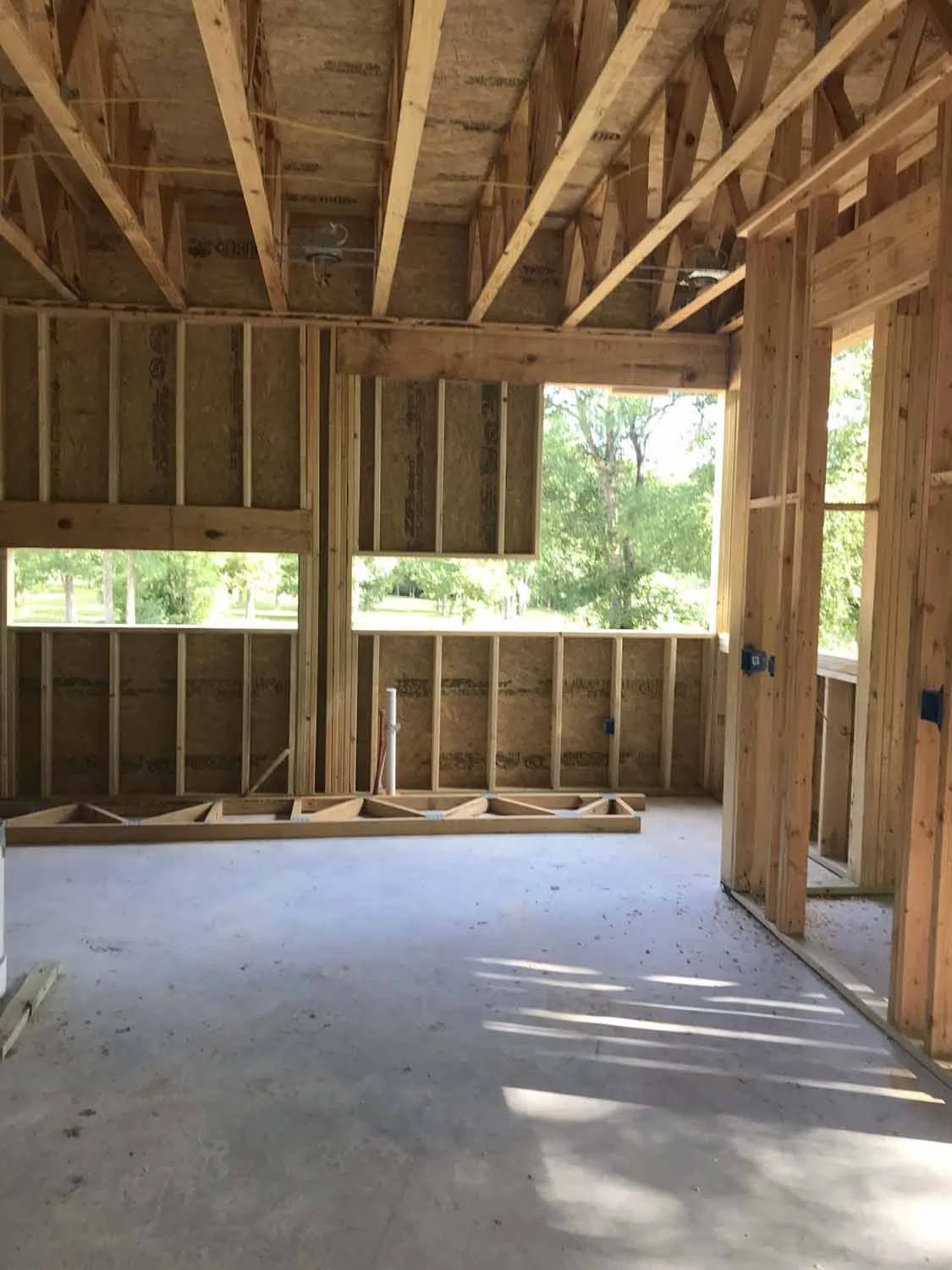 Interior of a house under construction, showing a wooden framing structure and large windows with a view of trees outside.