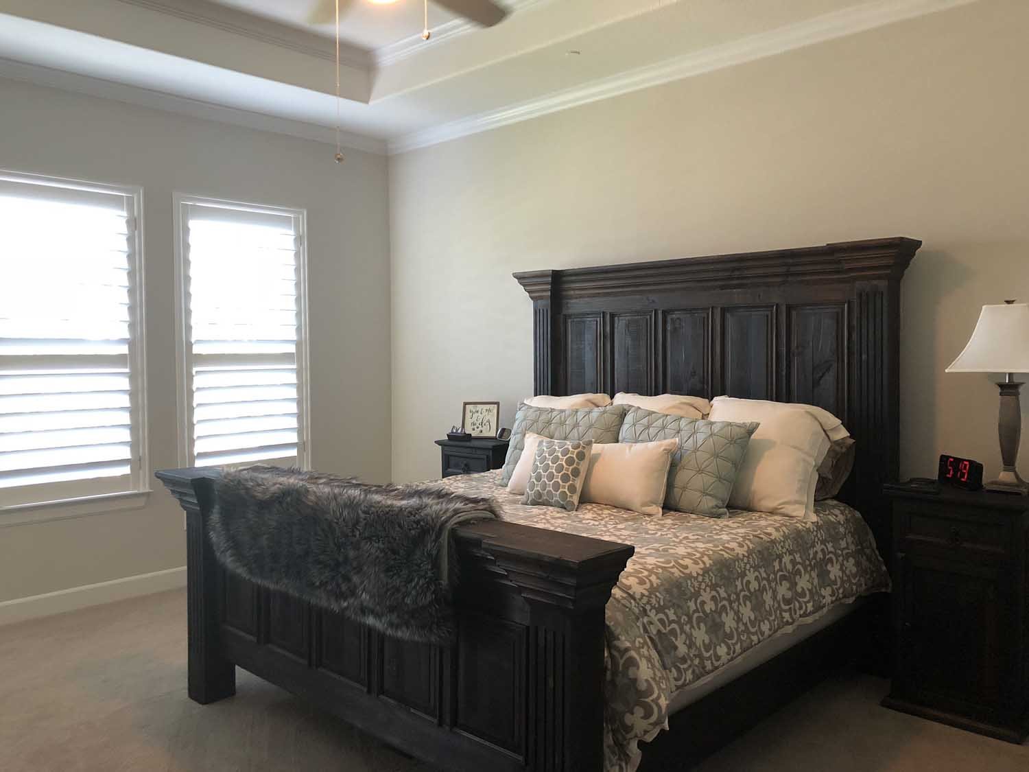 A bedroom with two windows with white blinds, a large dark wooden bed with decorative pillows, a fur throw, two nightstands with lamps, a digital clock, and a small framed picture.