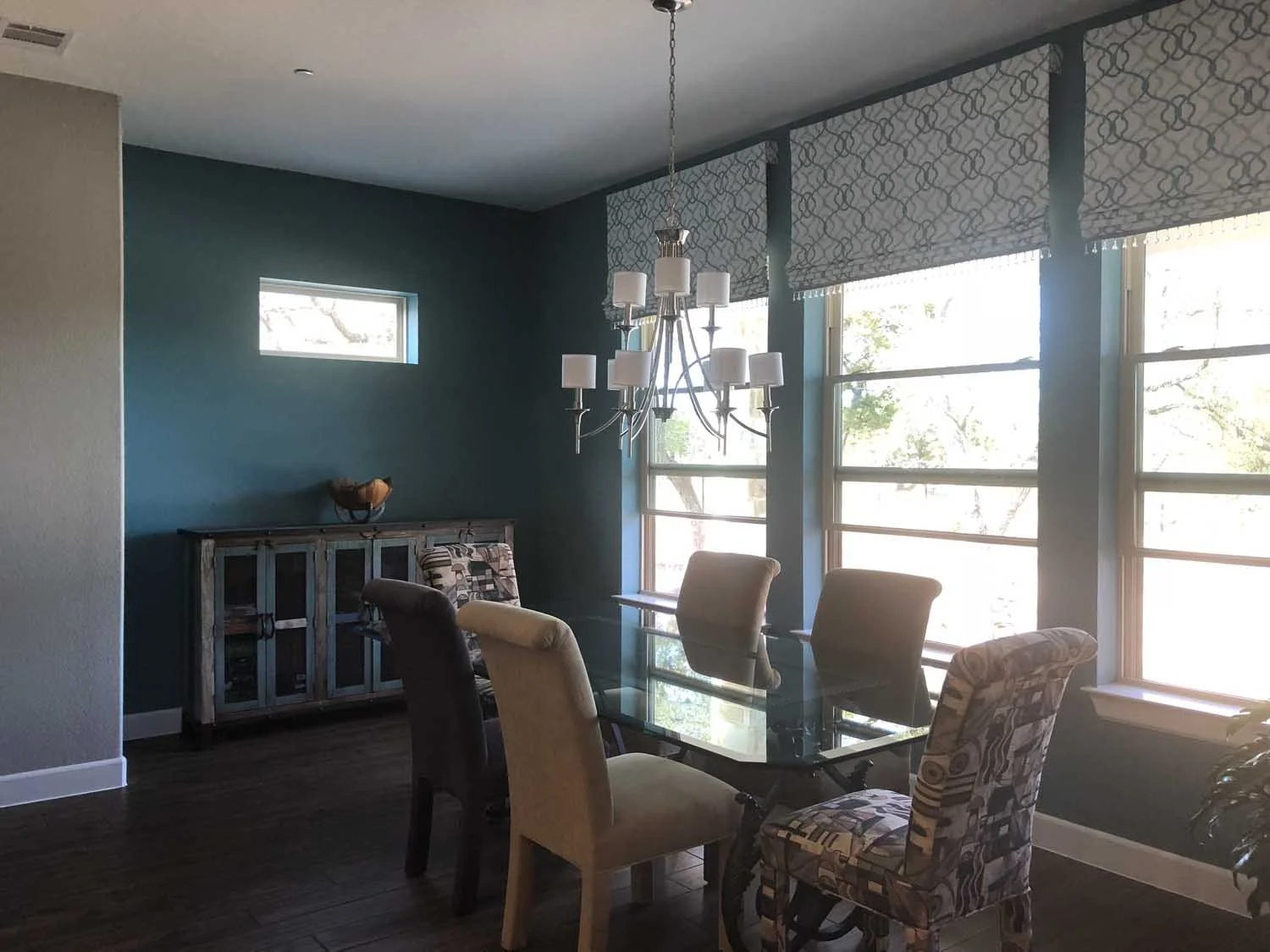 Dining room with a glass table, upholstered chairs, a dark wooden sideboard, and large windows with window shades letting in natural light.