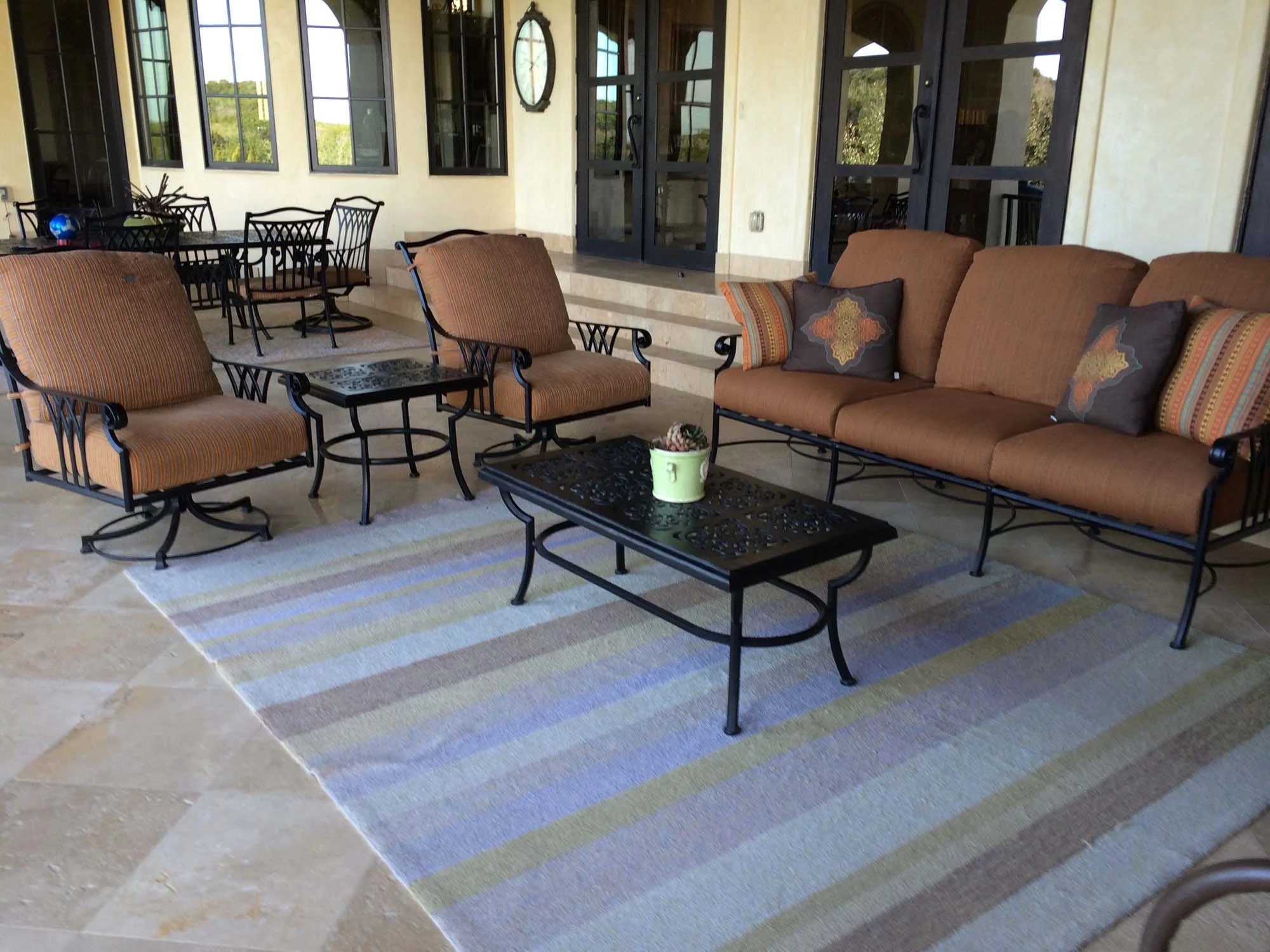 Patio furniture setup with a sofa, two armchairs, a coffee table, and additional chairs in the background on a tiled floor and a striped rug, with windows and a door behind.