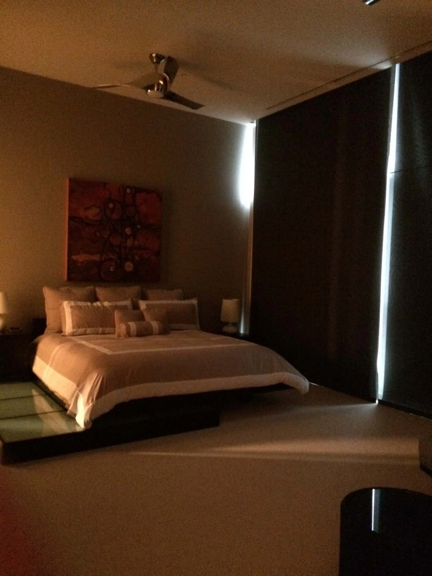 Bedroom with a bed, pillows, bedside lamps, a nightstand, abstract wall art, and closed blackout curtains with a small slit of light.