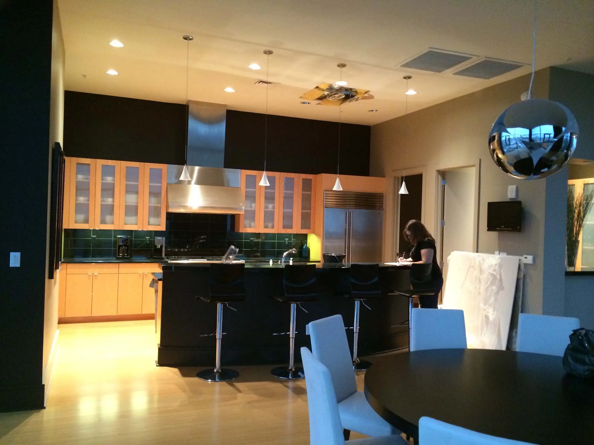Modern kitchen with wooden cabinets, black countertops, green tile backsplash, and a hallway with a woman writing at the counter.