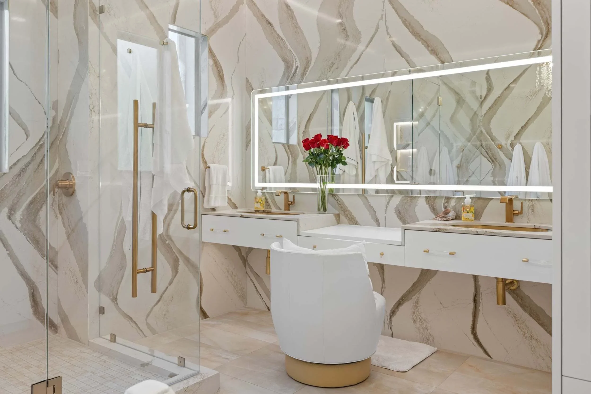 Modern bathroom with a large mirror, white cabinetry with gold handles, a white chair, and a glass shower with gold fixtures. A bouquet of red roses is on the countertop.