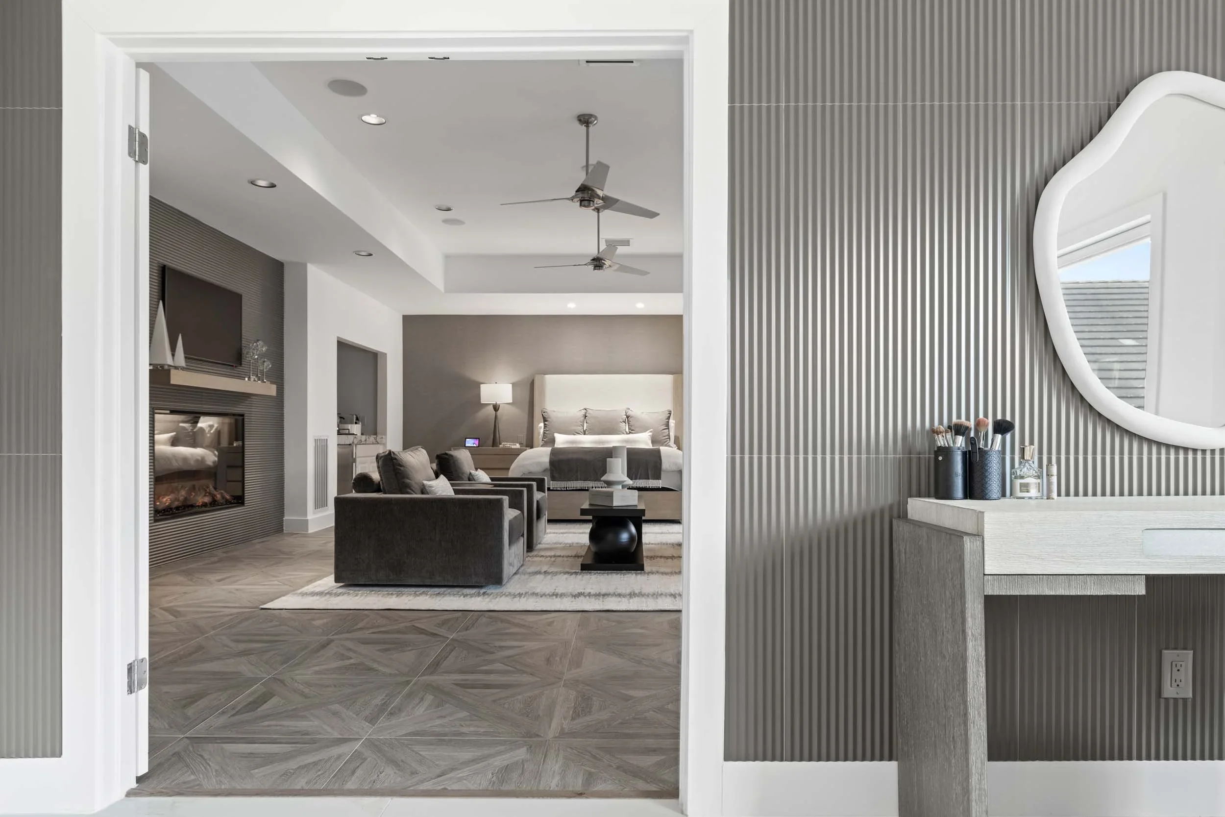 View into a modern, well-decorated bedroom with a sitting area and a fireplace, seen through an open doorway, with a vanity or dresser with makeup brushes and a mirror in the foreground.