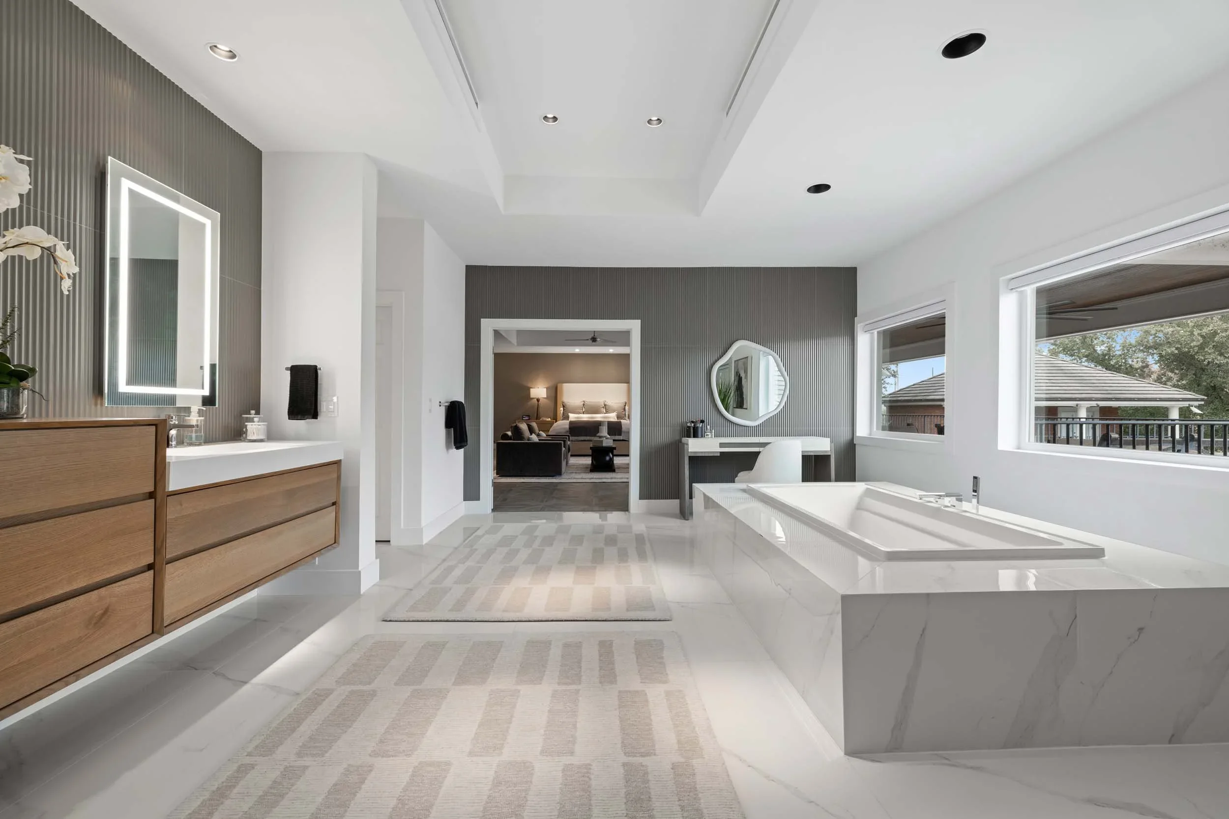 Modern bathroom with a white bathtub, marble tiles, wooden vanity, illuminated mirror, and large windows showing a view outside.