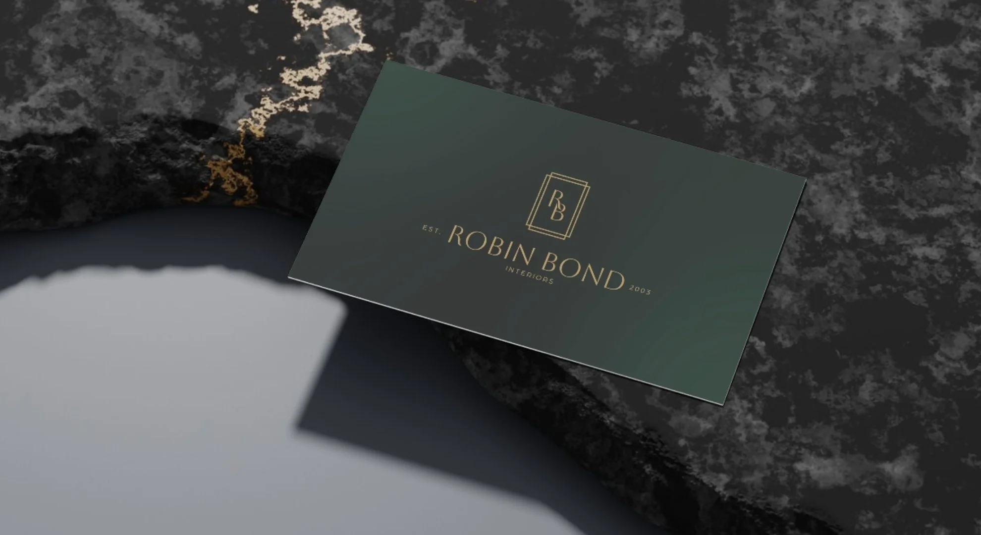 A business card for Robin Bond Interiors lies on a dark, textured surface with some gold accents. The card features a logo with the initials RB and the text 'Robin Bond Interiors' with 'EST 2003'.