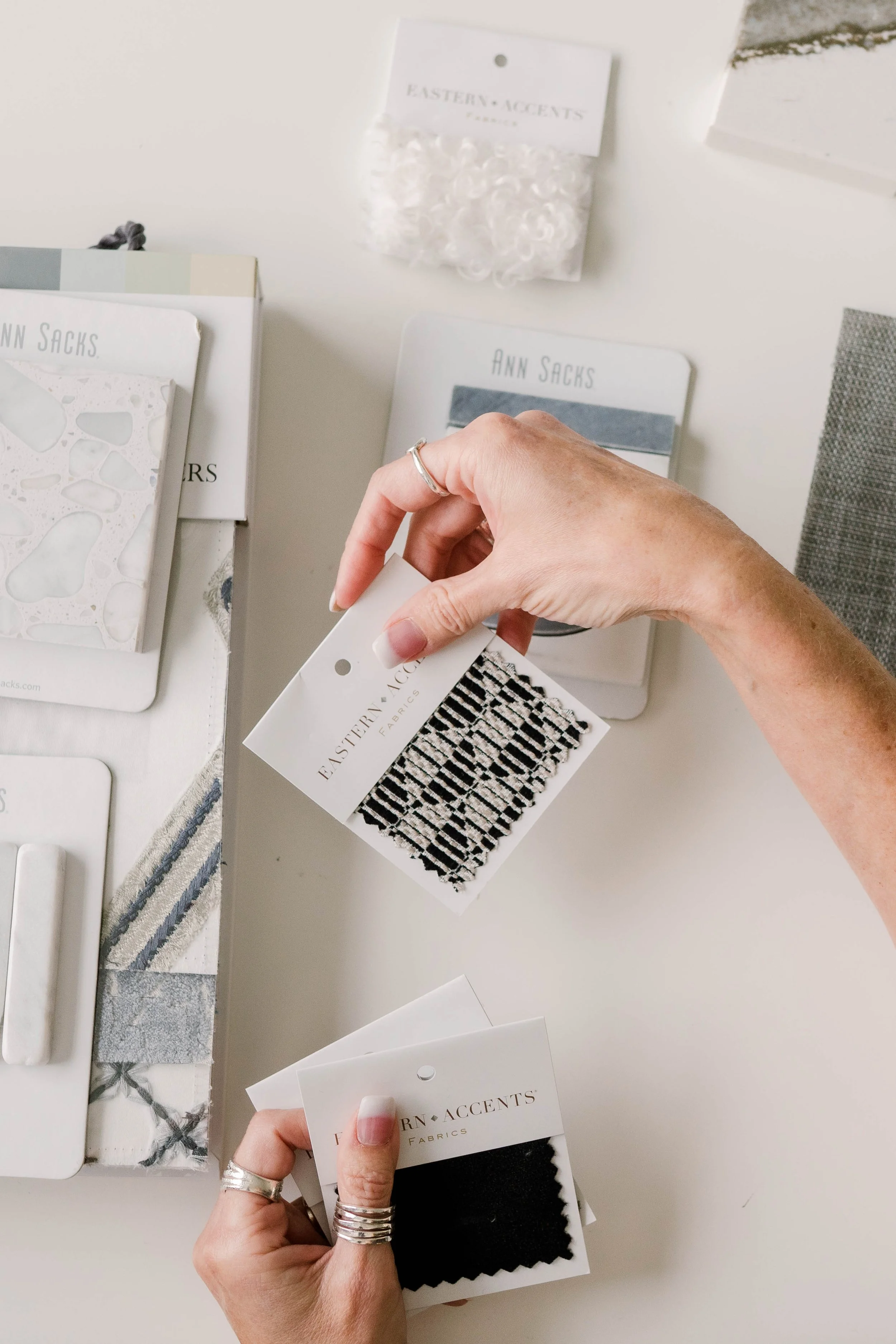 Hands holding fabric swatches over a white table with fabric design samples, patterns, and packaging labeled 'Eastern Accents' and 'Ann Sacks'.