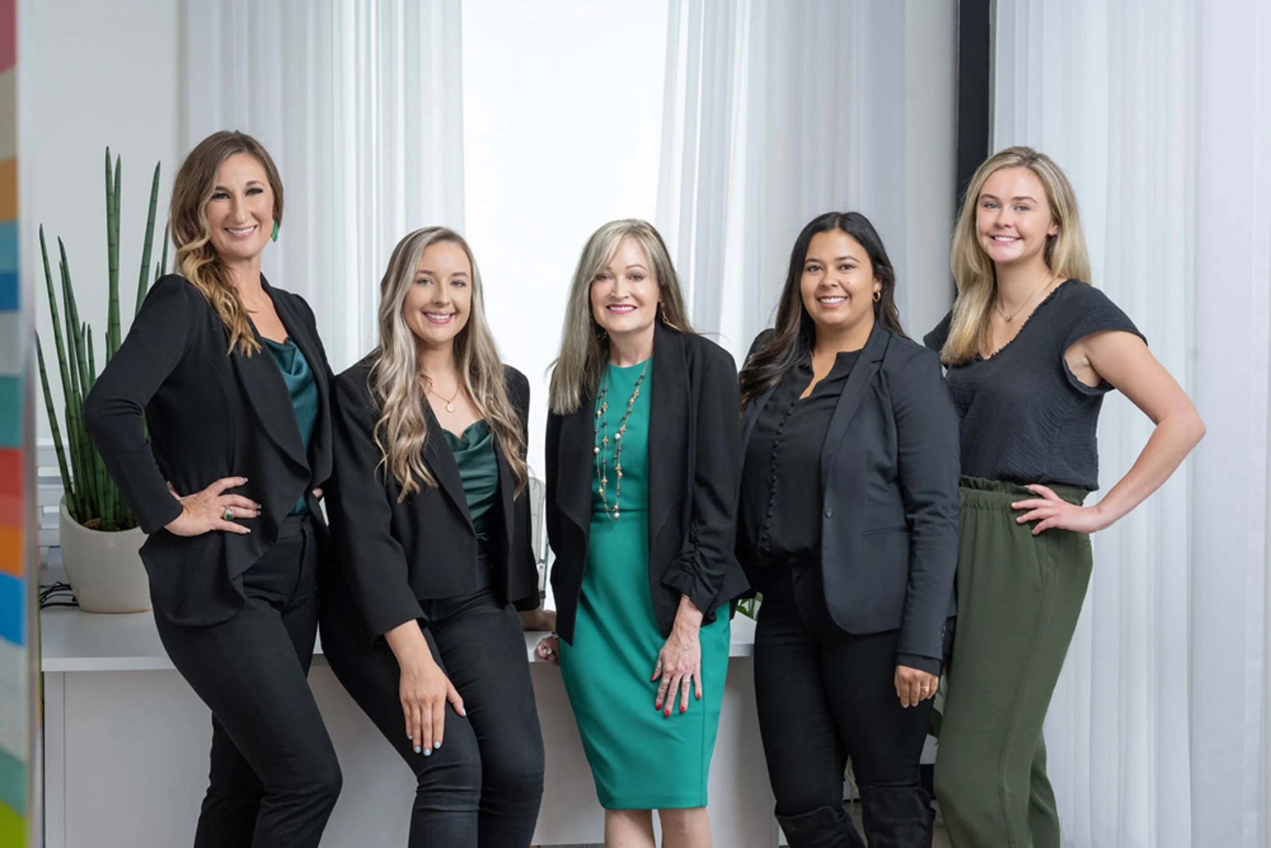 Group of five women dressed in business attire standing together indoors, smiling at the camera.