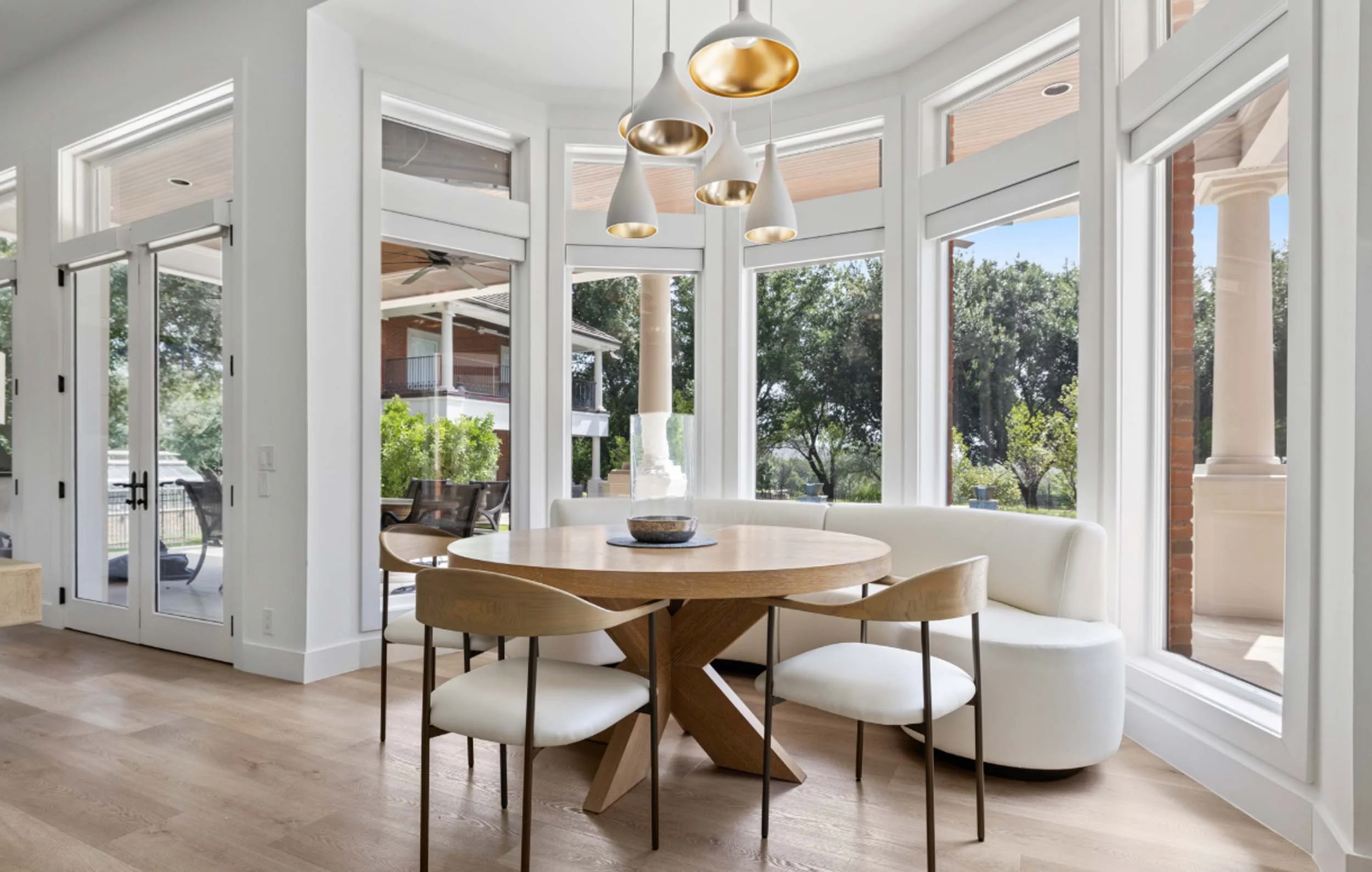Bright dining area with round wooden table, white modern chairs, curved bench, large windows, and contemporary pendant lights, designed by Robin Bond Interiors.