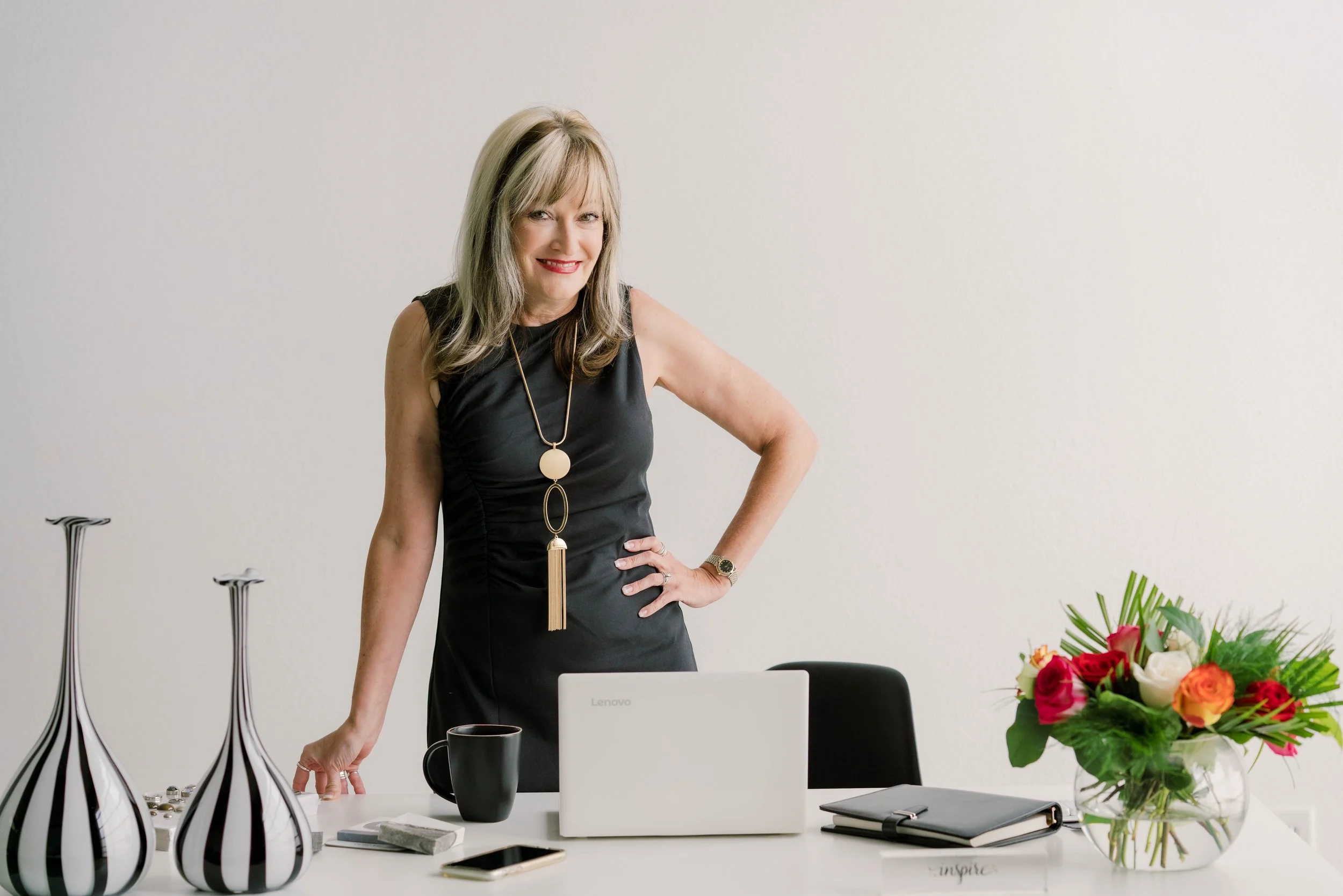 A woman in a black dress standing behind a white desk with a laptop, a black mug, a bouquet of multicolored flowers, and decorative vases.