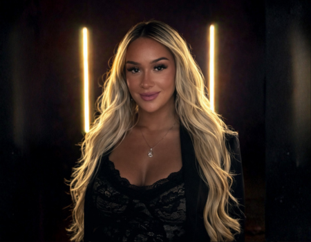 A woman with long, wavy blonde hair smiling at the camera with illuminated vertical lights in the background.