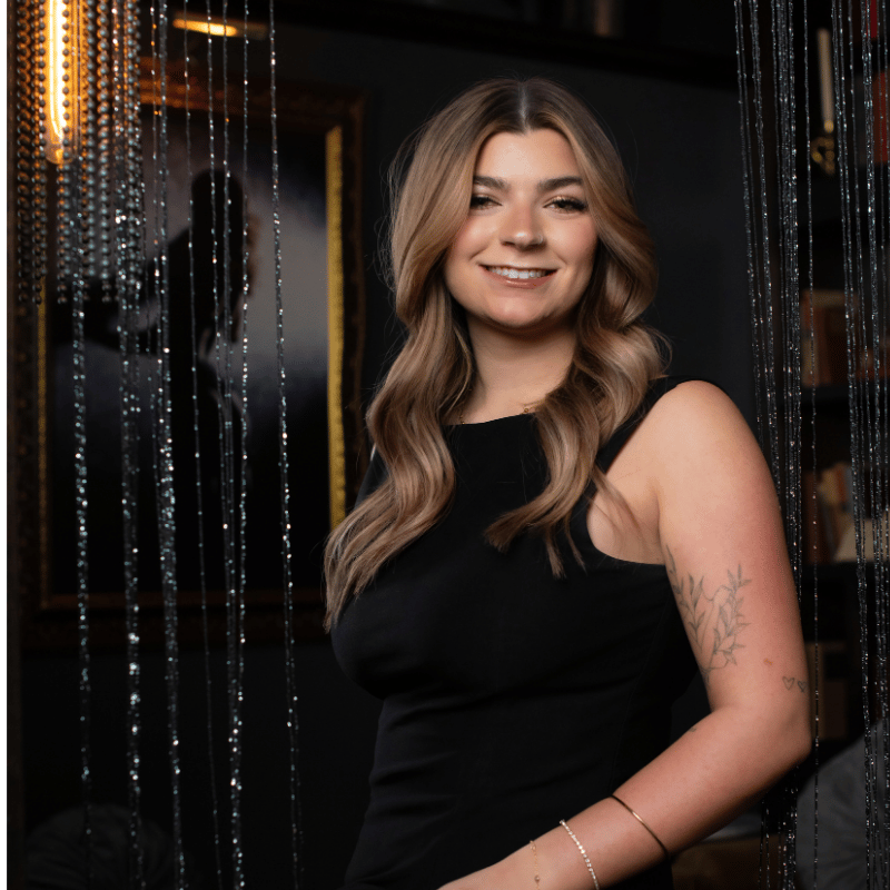 Portrait of a young woman with long, wavy, reddish-brown hair, smiling softly, wearing layered necklaces, with neon orange lights in the background.