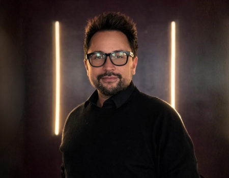 Portrait of a man wearing glasses, with short dark hair and a beard, in front of a dark background with vertical orange light streaks.