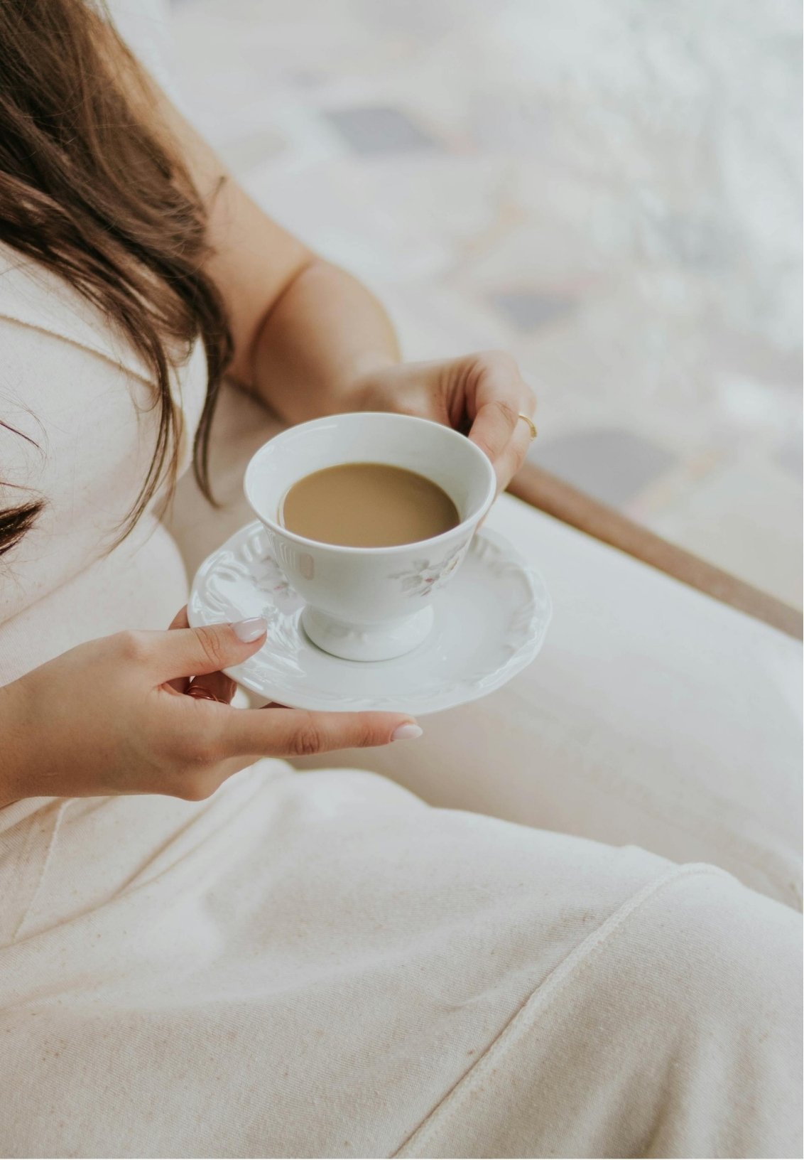 A woman in a beige outfit is holding a white porcelain teacup filled with tea or coffee and a matching saucer, sitting on a light-colored surface.