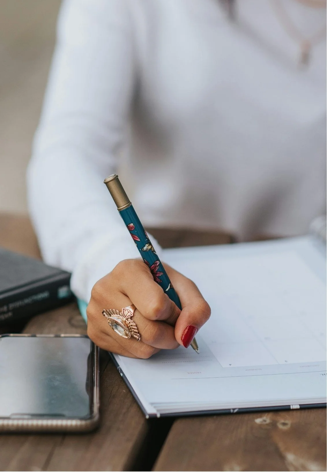 A woman writing with a pen in a planner or notebook, wearing a large decorative ring and red nail polish, sitting at a wooden table with a smartphone and a book nearby.
