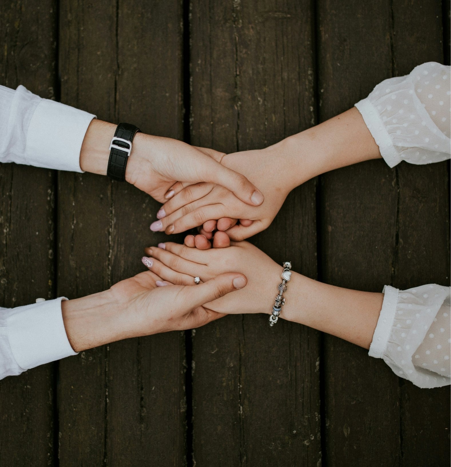 Four hands placed together in the center of a dark wooden surface with two wearing bracelets and one wearing a ring.