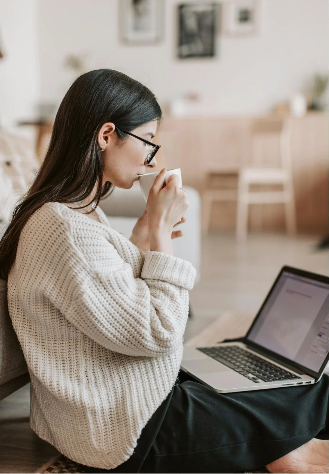A woman with glasses sitting on the floor, drinking from a white mug, with a laptop open on her lap in a cozy living room.