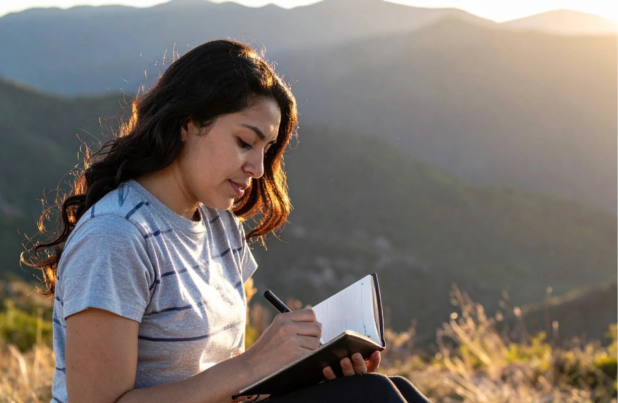 A woman with dark wavy hair, wearing a gray striped t-shirt, sitting outdoors on a hillside at sunset, writing in a notebook.