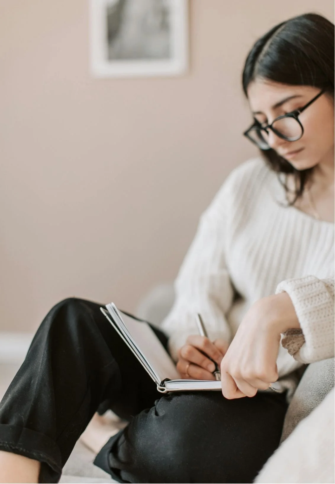 A woman with black hair, glasses, and a cream-colored sweater sitting on the floor, writing in a notebook.