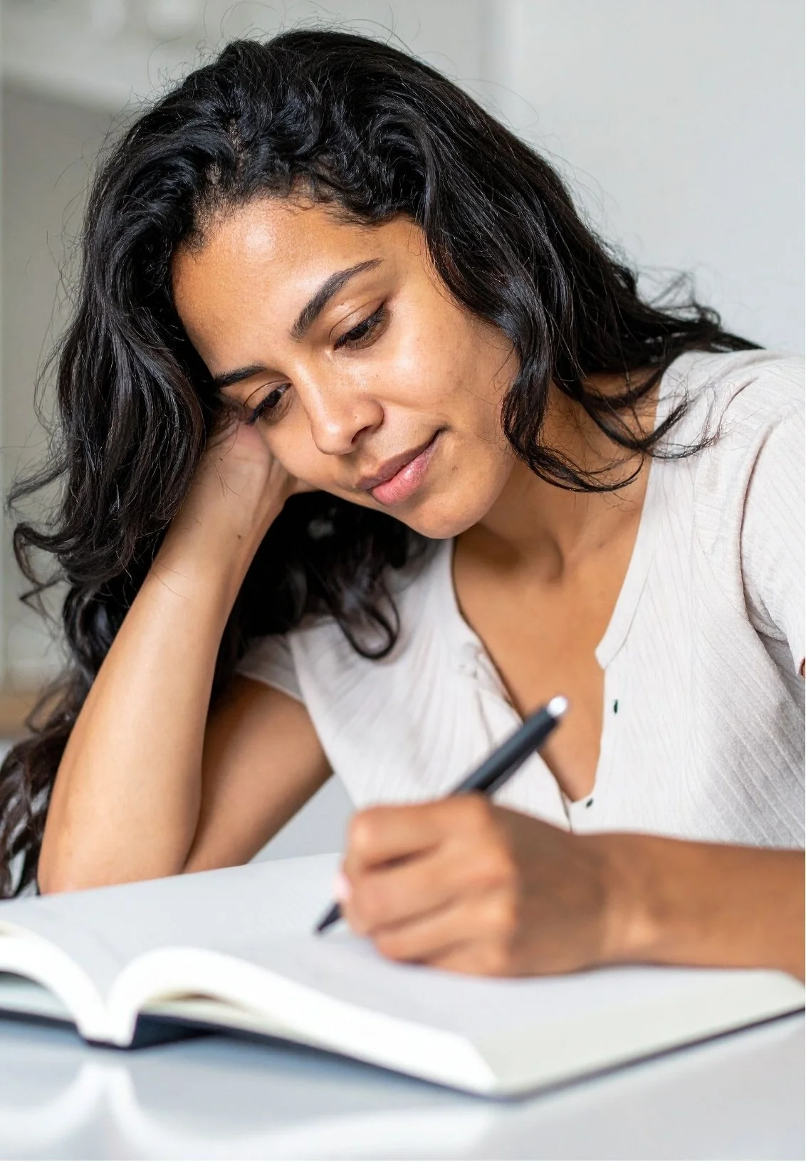 A woman with curly black hair writing in a notebook, resting her head on her left hand with a thoughtful expression.