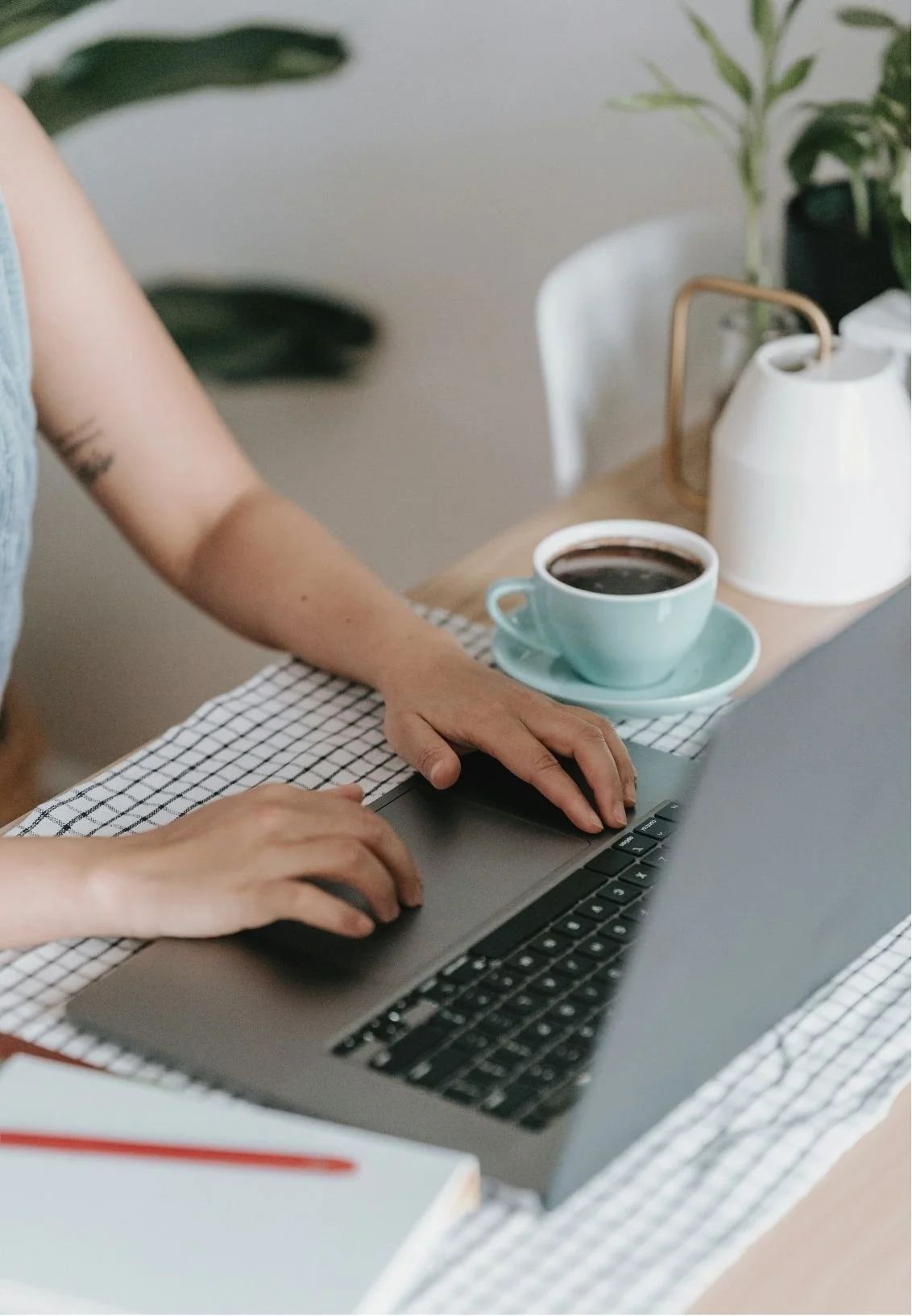 A person typing on a laptop at a dining table with a cup of coffee, a notebook with a red apple, and a white pitcher in the background.