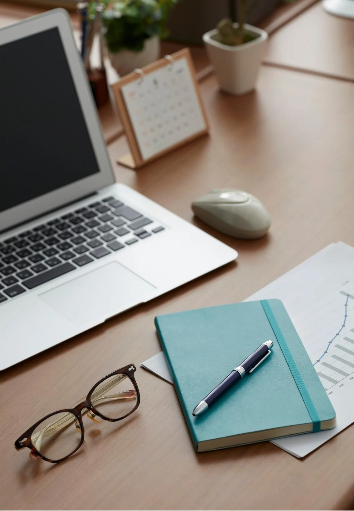 A tidy workspace with a laptop, glasses, a closed teal notebook with a pen on top, a mouse, some papers with graphs, a calendar, a potted plant, and a small container on a wooden desk.