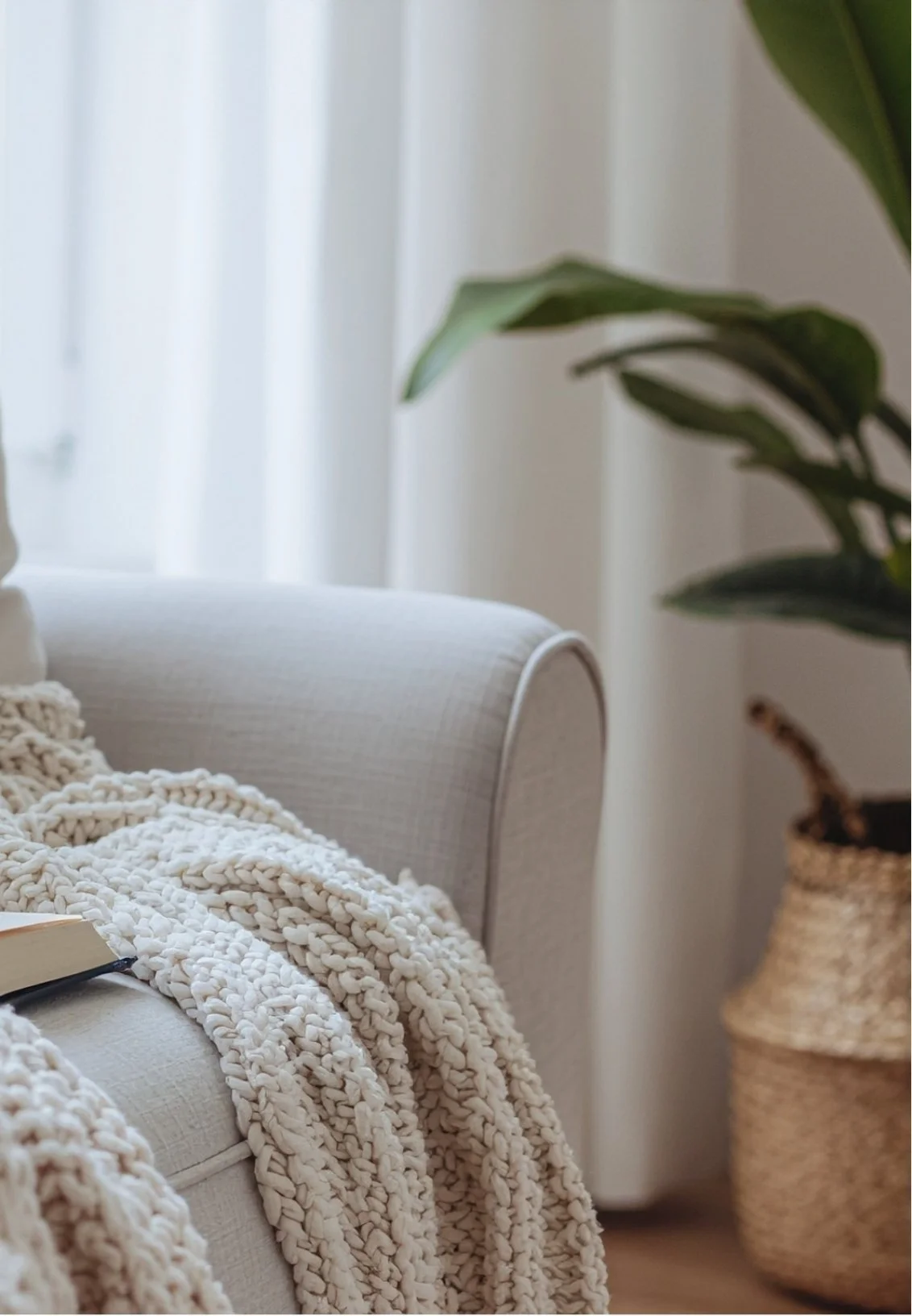 A cozy living room scene with a beige upholstered armchair, a chunky cream knit blanket, a small open book, and a ceramic planter with a large green leafy plant in the background.