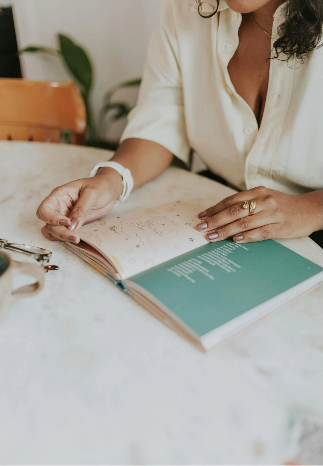 A woman reading an open book with handwritten notes and drawings inside, on a cream-colored table, with a pair of glasses nearby.