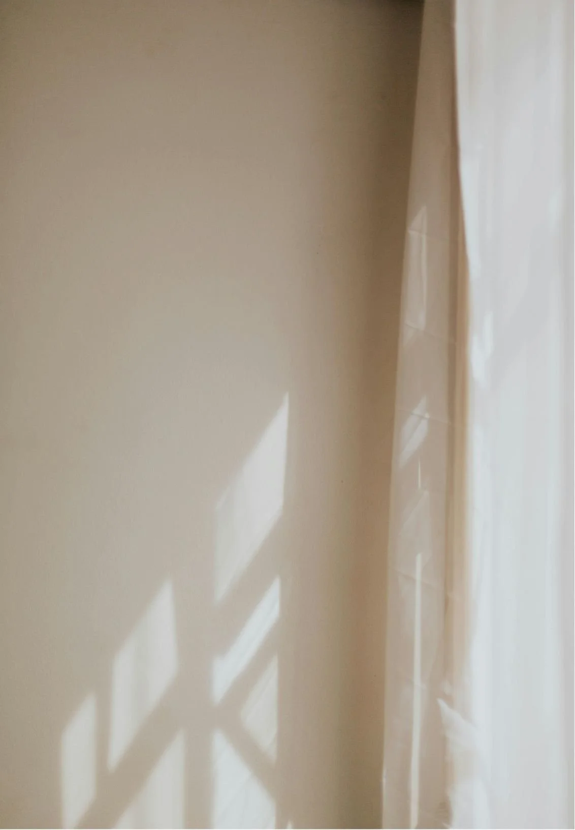 Sunlight casting shadows of window panes onto a light-colored wall with a partially visible curtain.