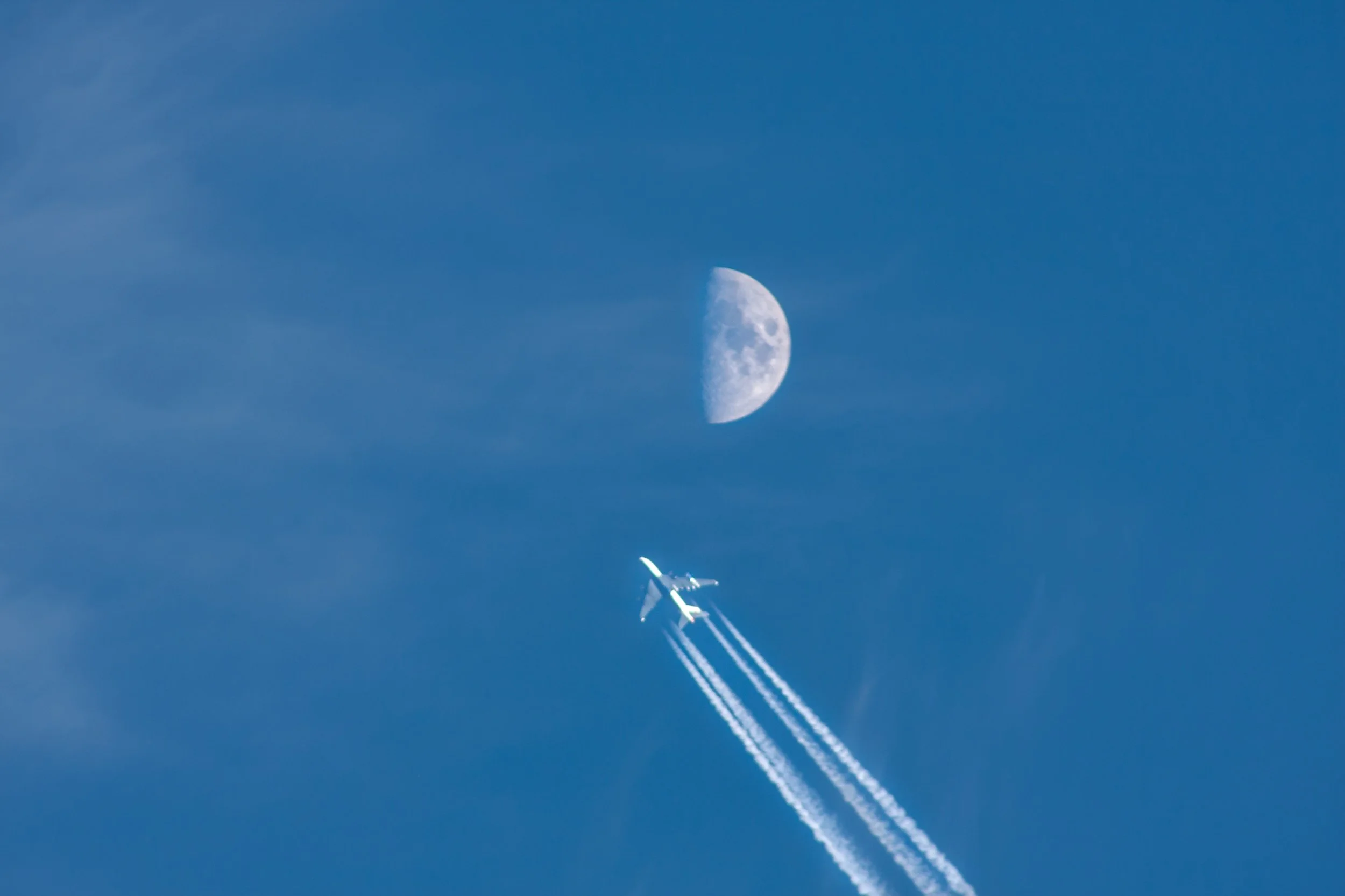 An aircraft leaves white contrails beneath the daytime moon.