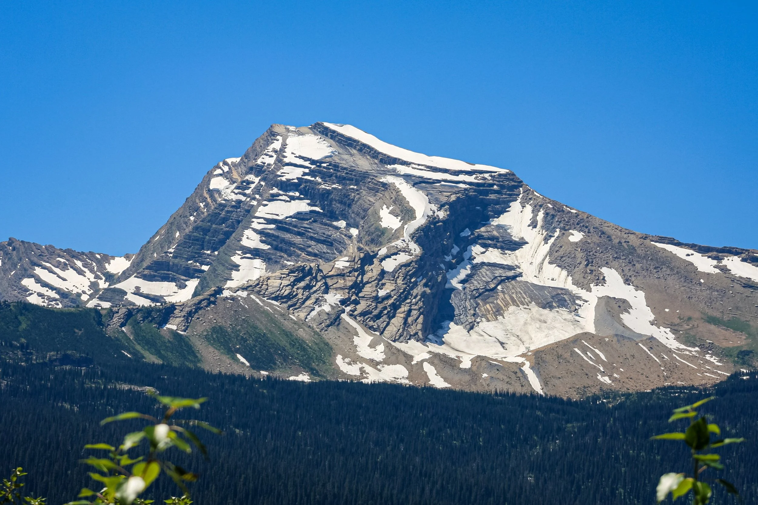 A snow-streaked alpine peak rises above a forested slope.