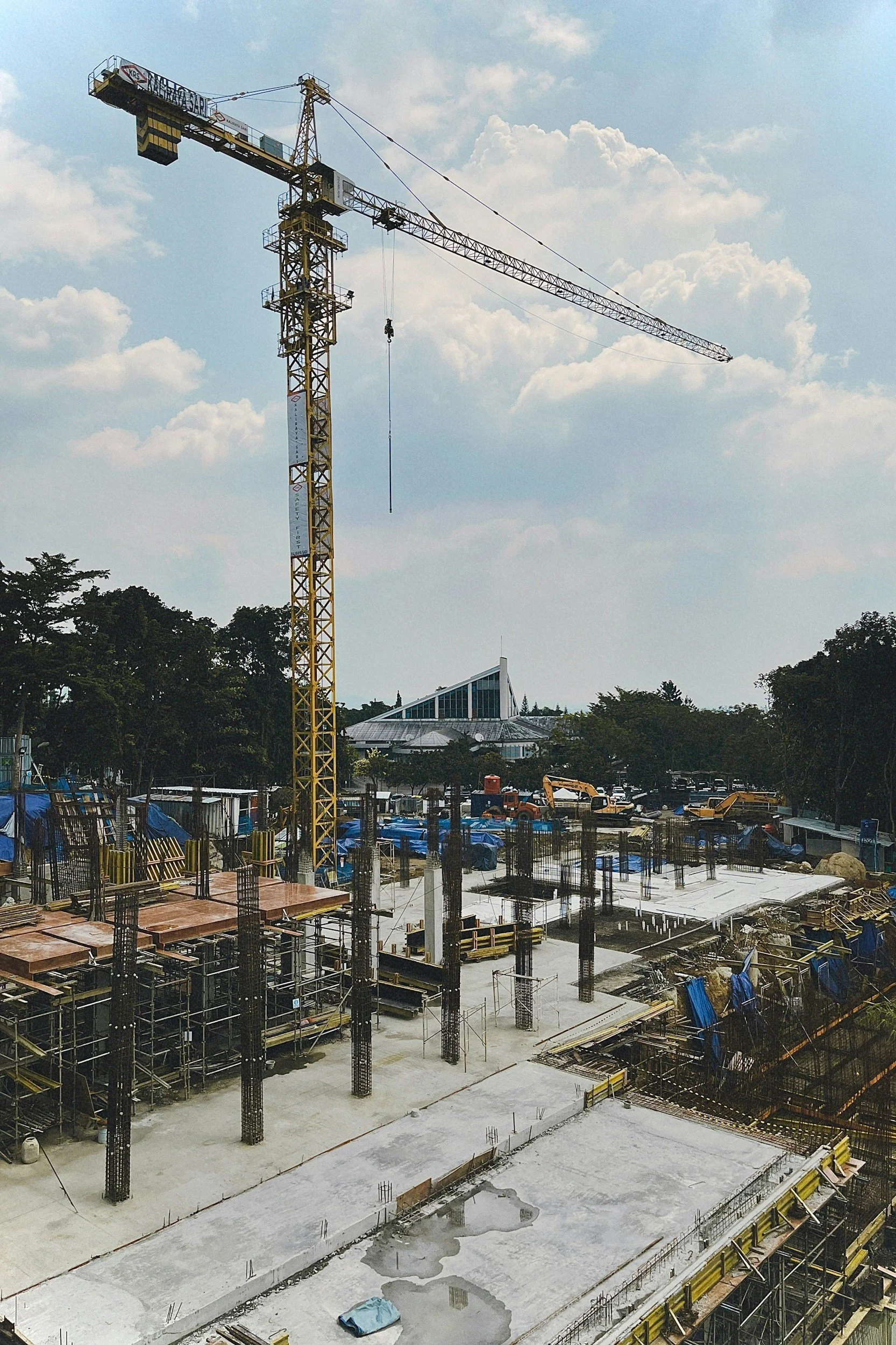 Construction site with a yellow tower crane, concrete foundation, and construction equipment, with trees and a modern building in the background.