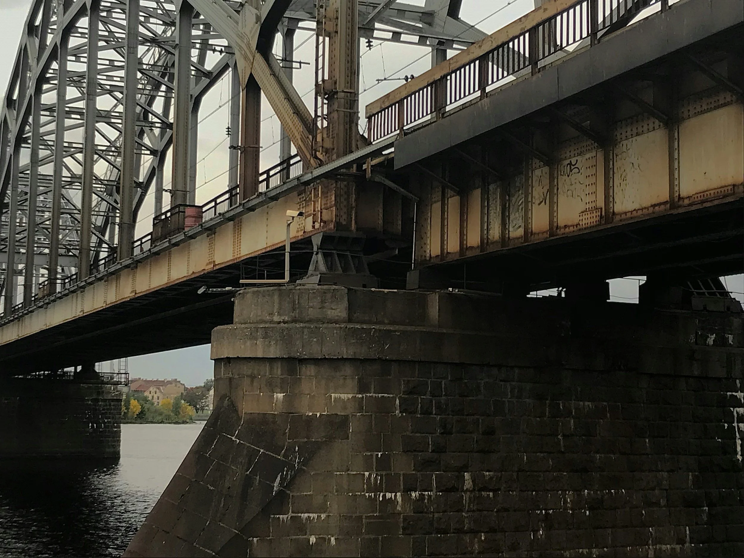 Close-up view of an old bridge with rusty yellow steel beams, metal rails, and a stone foundation over a body of water, with houses and trees in the background under an overcast sky.