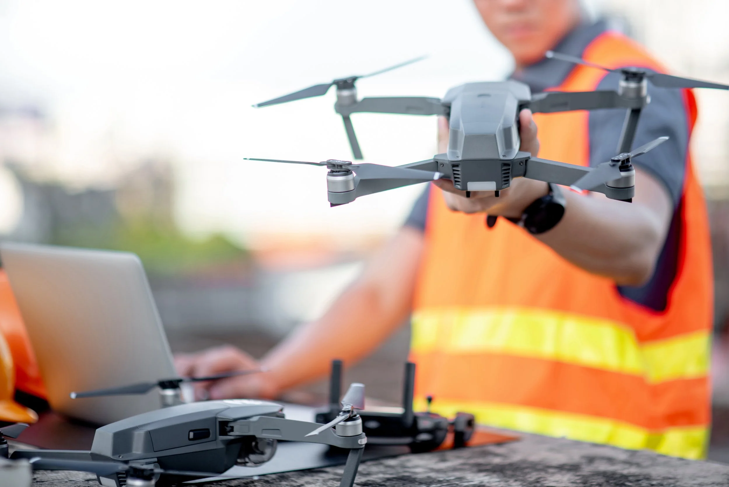 Person wearing an orange safety vest holding a drone outdoors, with a laptop nearby.