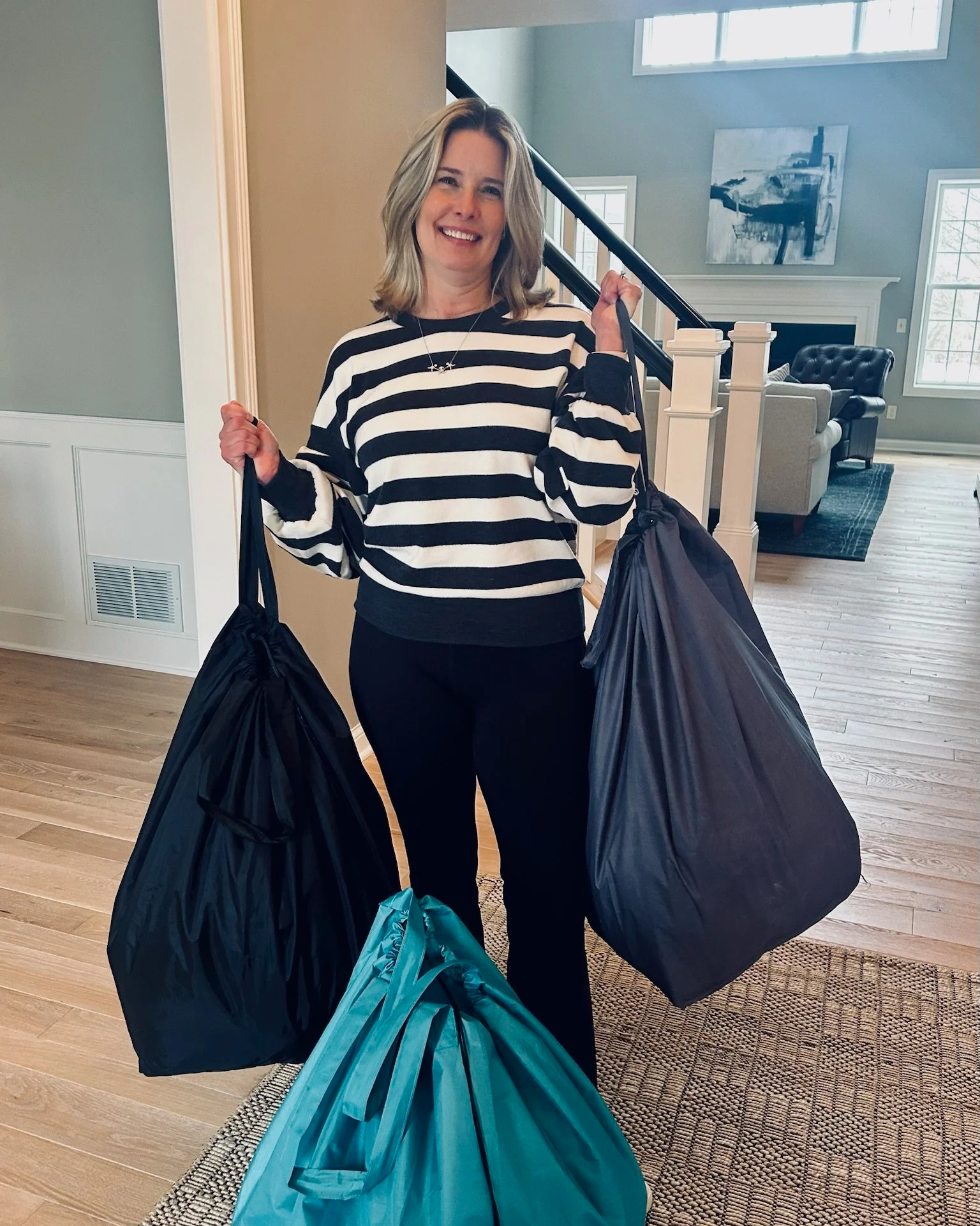 A smiling woman with shoulder-length blonde hair holding three large garbage bags inside a home with wooden floors, a staircase, and a living room in the background.