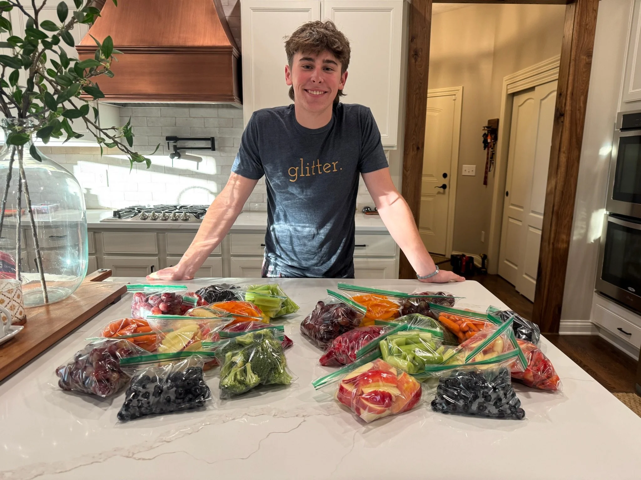 A young man with curly hair smiling and leaning on a kitchen counter covered with sealed plastic bags of assorted cut fruits and vegetables, including grapes, blueberries, blackberries, strawberries, celery, carrots, broccoli, and peach slices.