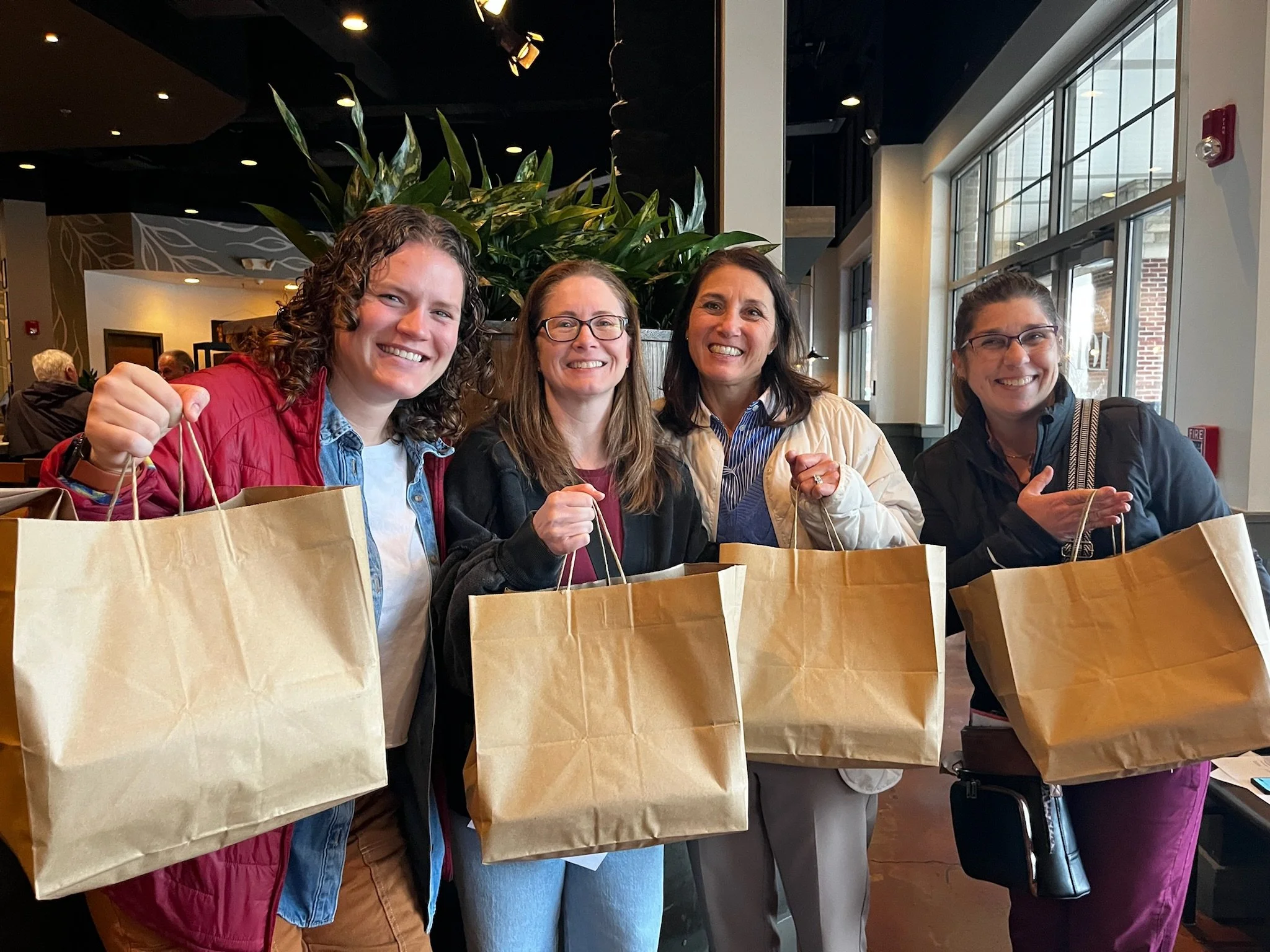 Four women standing inside a cafe, smiling at the camera, each holding a large brown paper shopping bag.