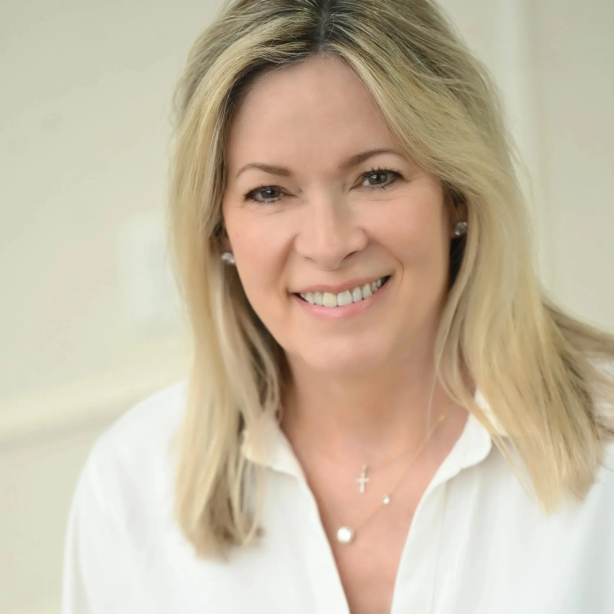 A smiling woman with blonde hair and earrings, wearing a white shirt and layered necklaces, in a light-colored indoor setting.