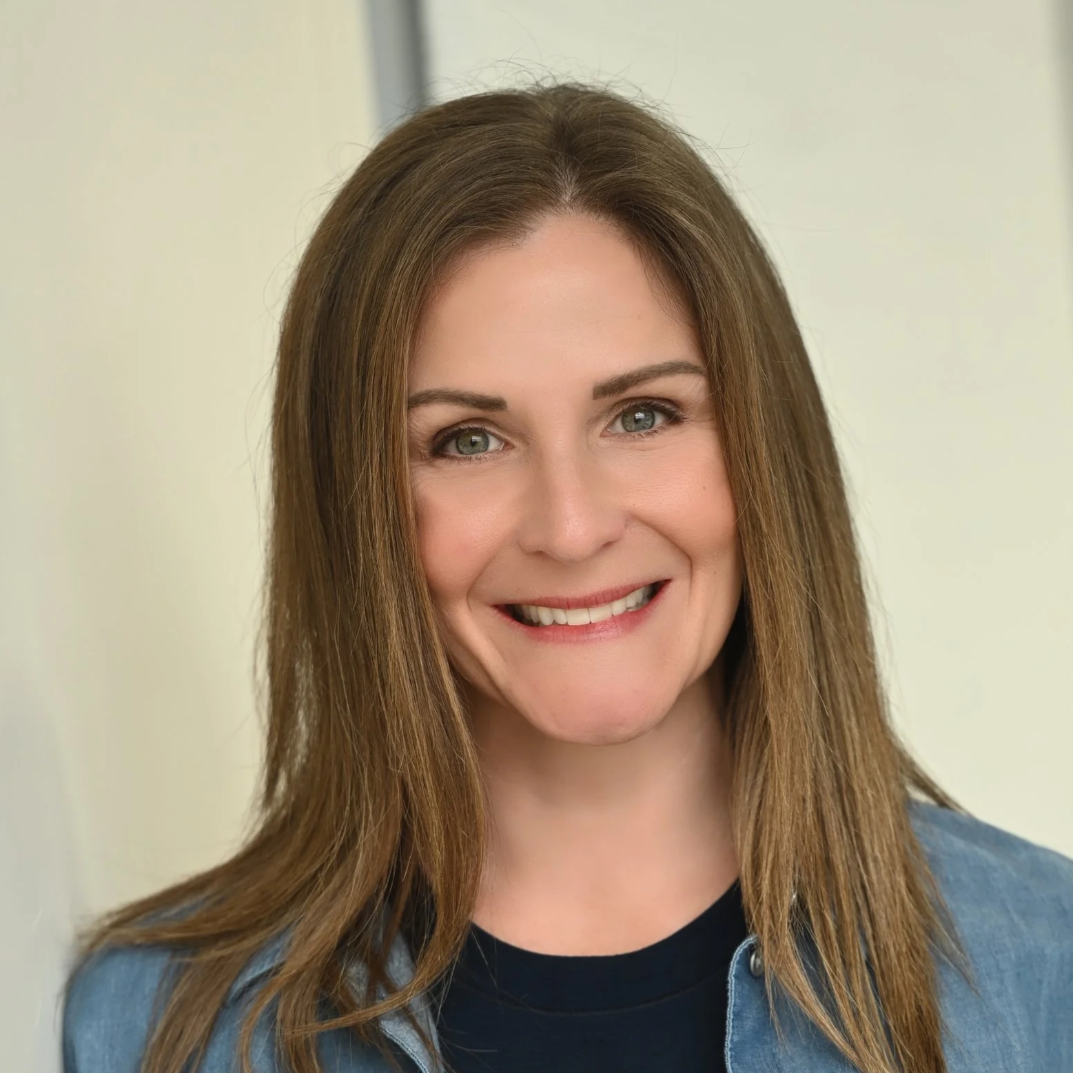 Close-up of a woman with shoulder-length brown hair, blue eyes, and a friendly smile, wearing a black shirt and a blue denim jacket against a plain background.
