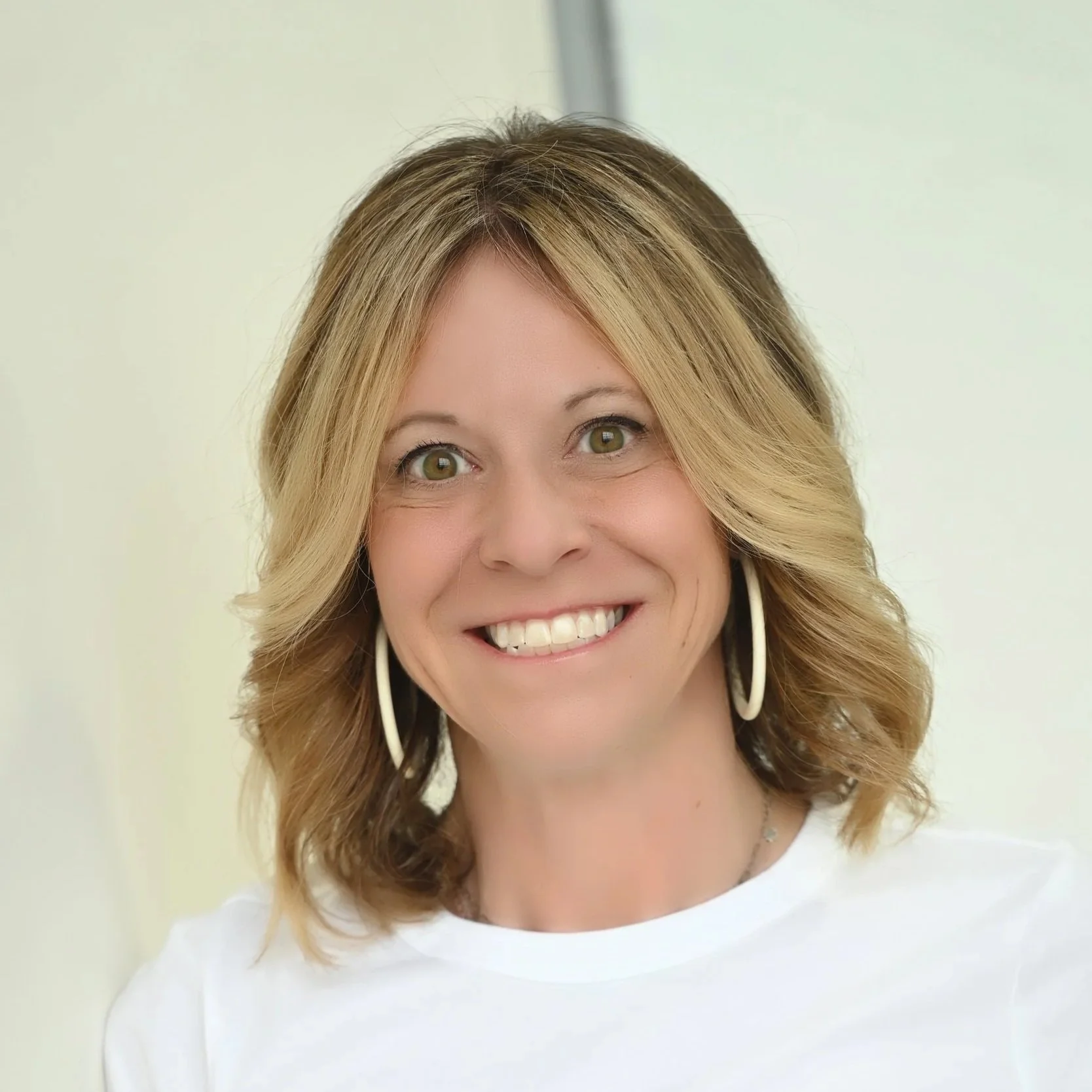 A smiling woman with shoulder-length light brown hair and green eyes, wearing white earrings and a white top, standing indoors with a neutral background.