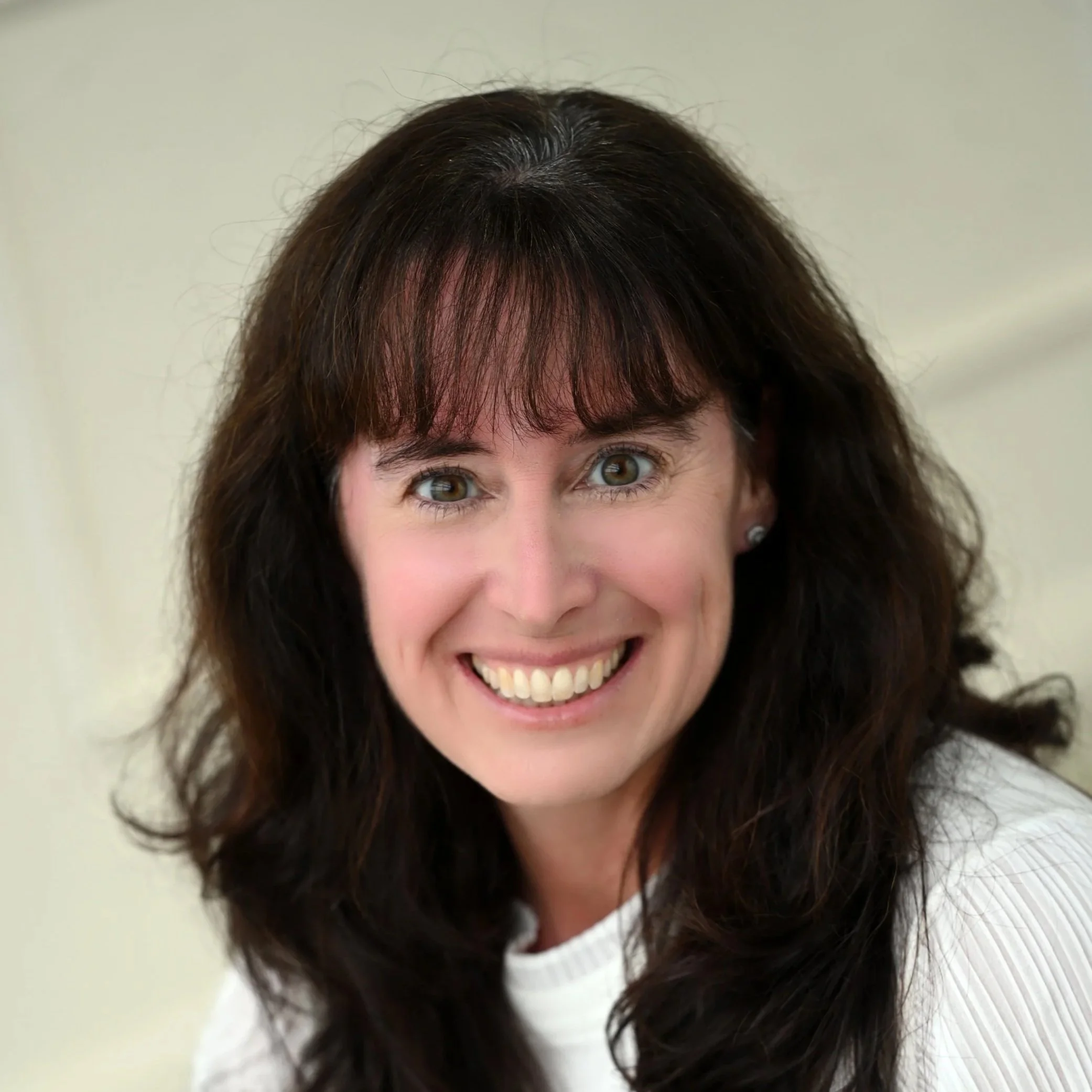 A woman with dark wavy hair, smiling, wearing a white top, against a light background.