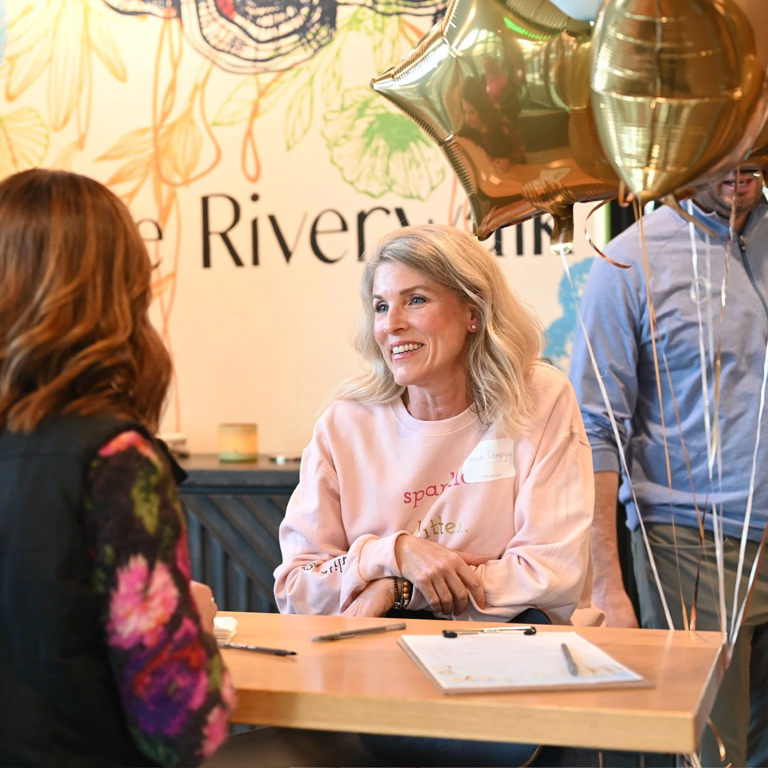 A woman with shoulder-length blonde hair smiling and talking to another woman with reddish-brown hair at a table with pens and paper, in a decorated room with balloons and a sign that says "The Riverview" in the background.