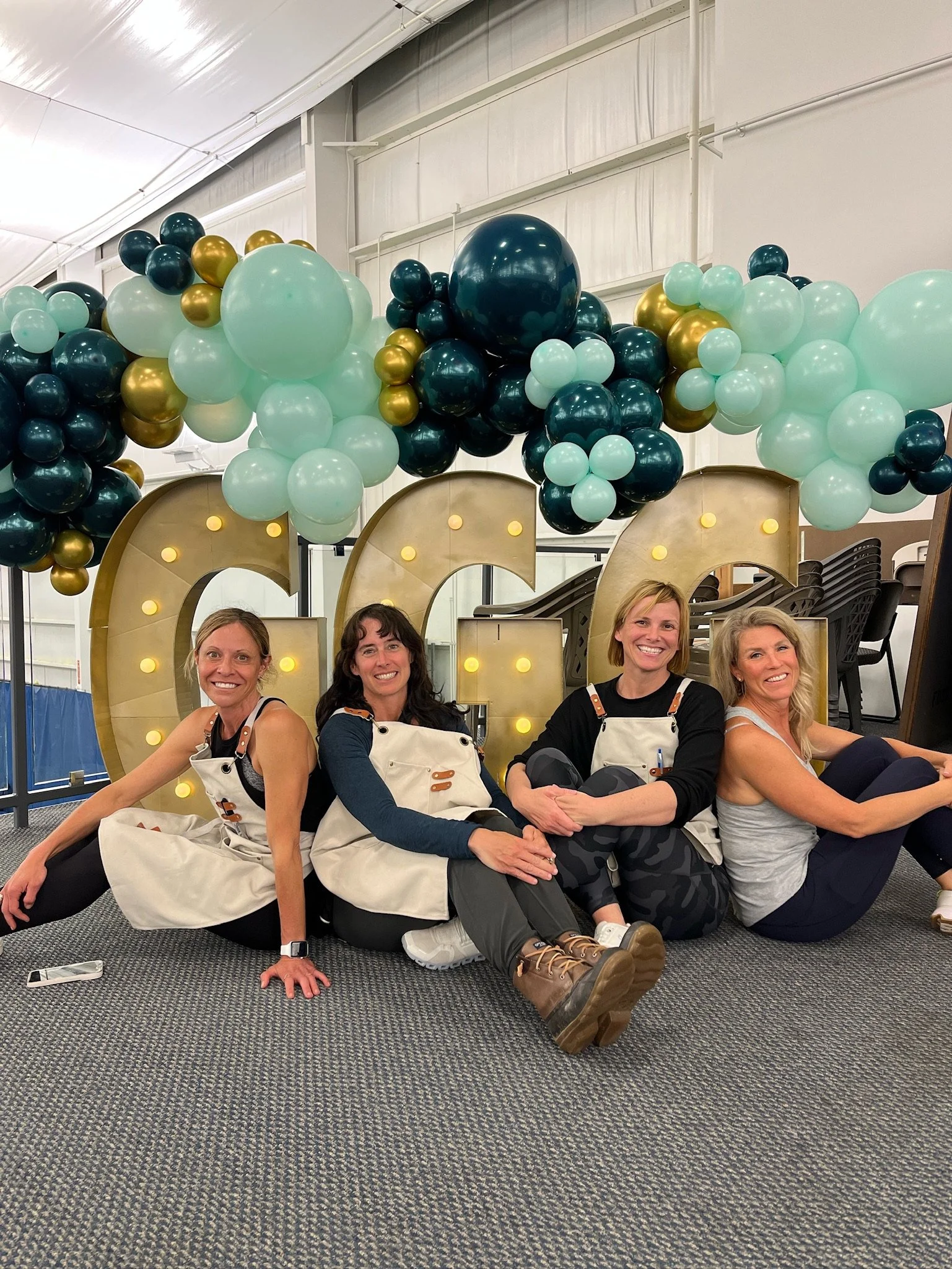 Four women sitting on the floor in front of large illuminated letters spelling 'GIRL,' with balloons in shades of teal, teal, and gold overhead, inside a large indoor space.