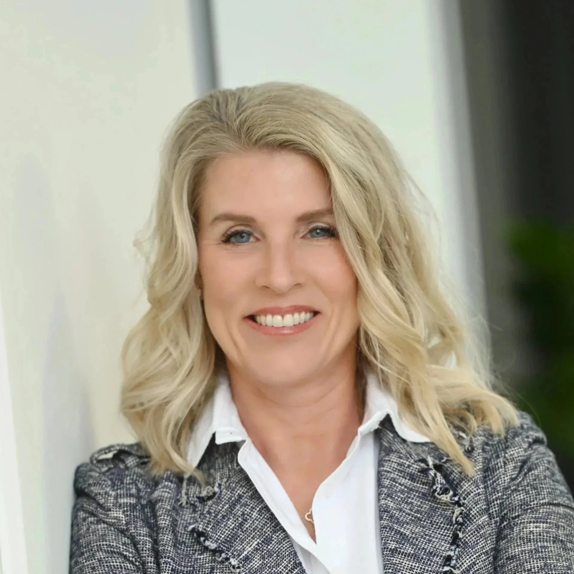 A smiling woman with blonde wavy hair, wearing a white shirt and a gray patterned blazer, standing indoors near a light-colored wall.